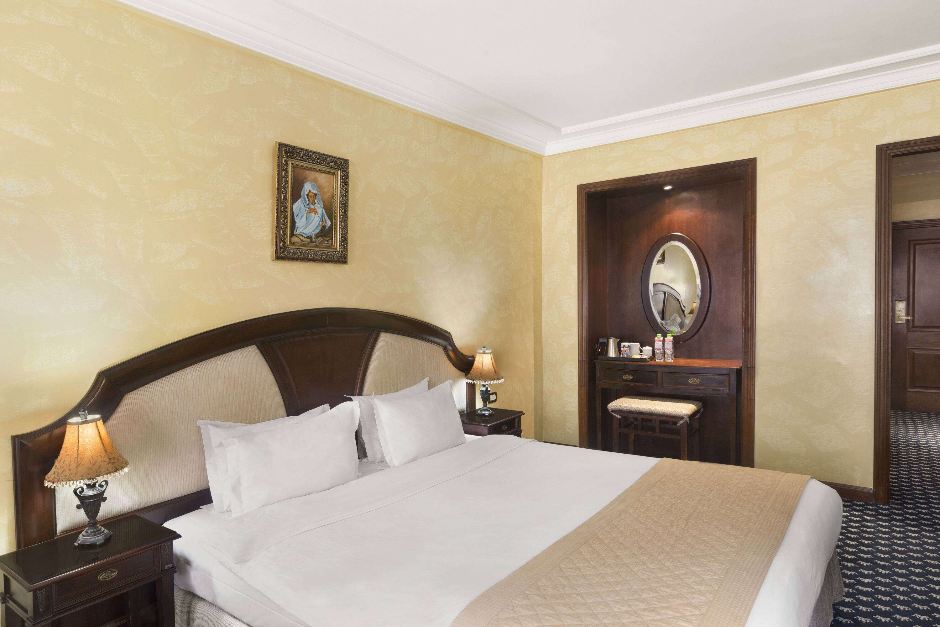 Habitación de hotel con una cama grande, una mesita de noche con lámpara, cabecero de madera, una pequeña mesa con espejo y un cuadro en la pared.