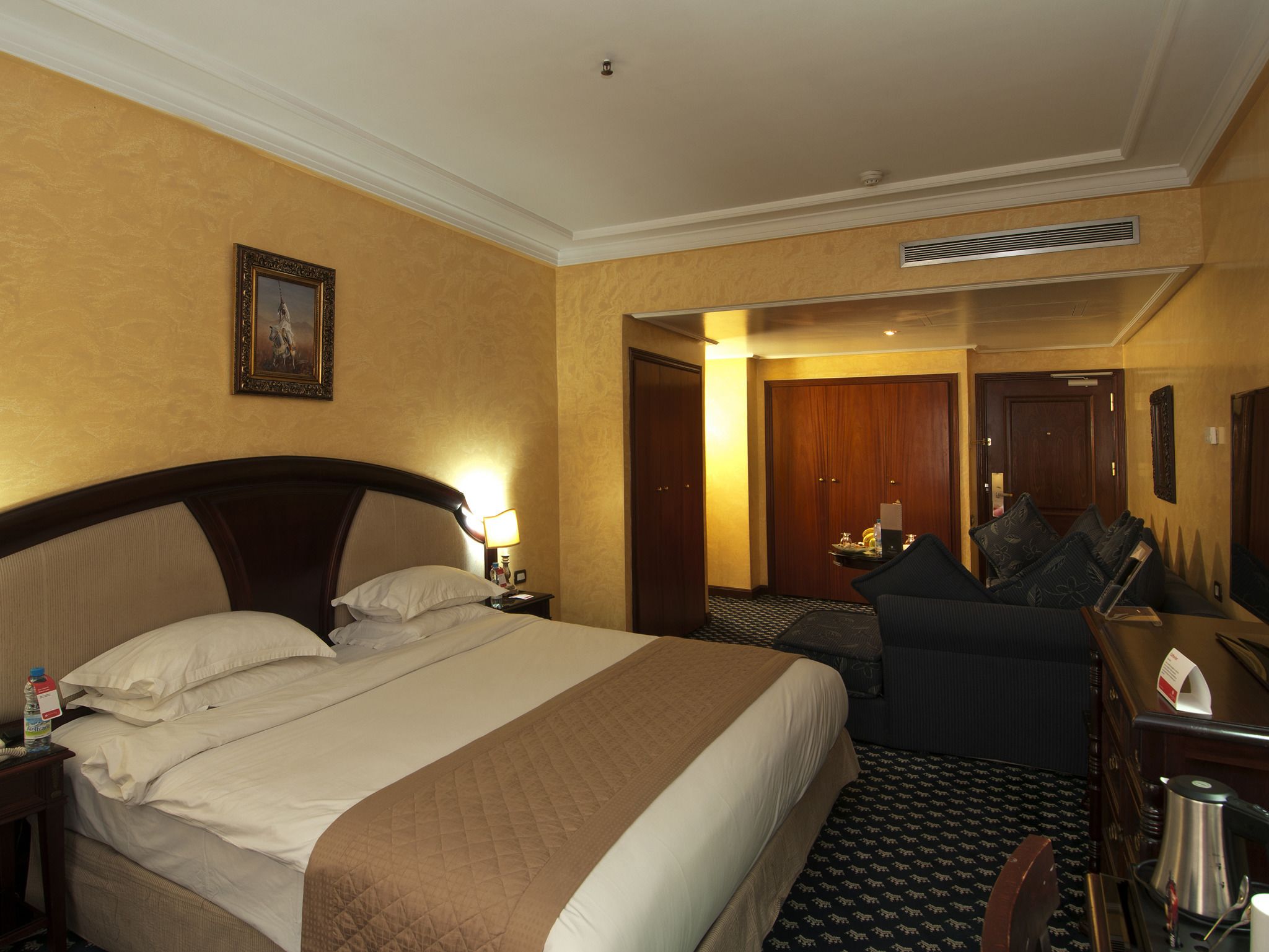 A hotel room with a large bed, a desk with a chair, a desk lamp, a framed picture on the wall, and a door leading to another room.