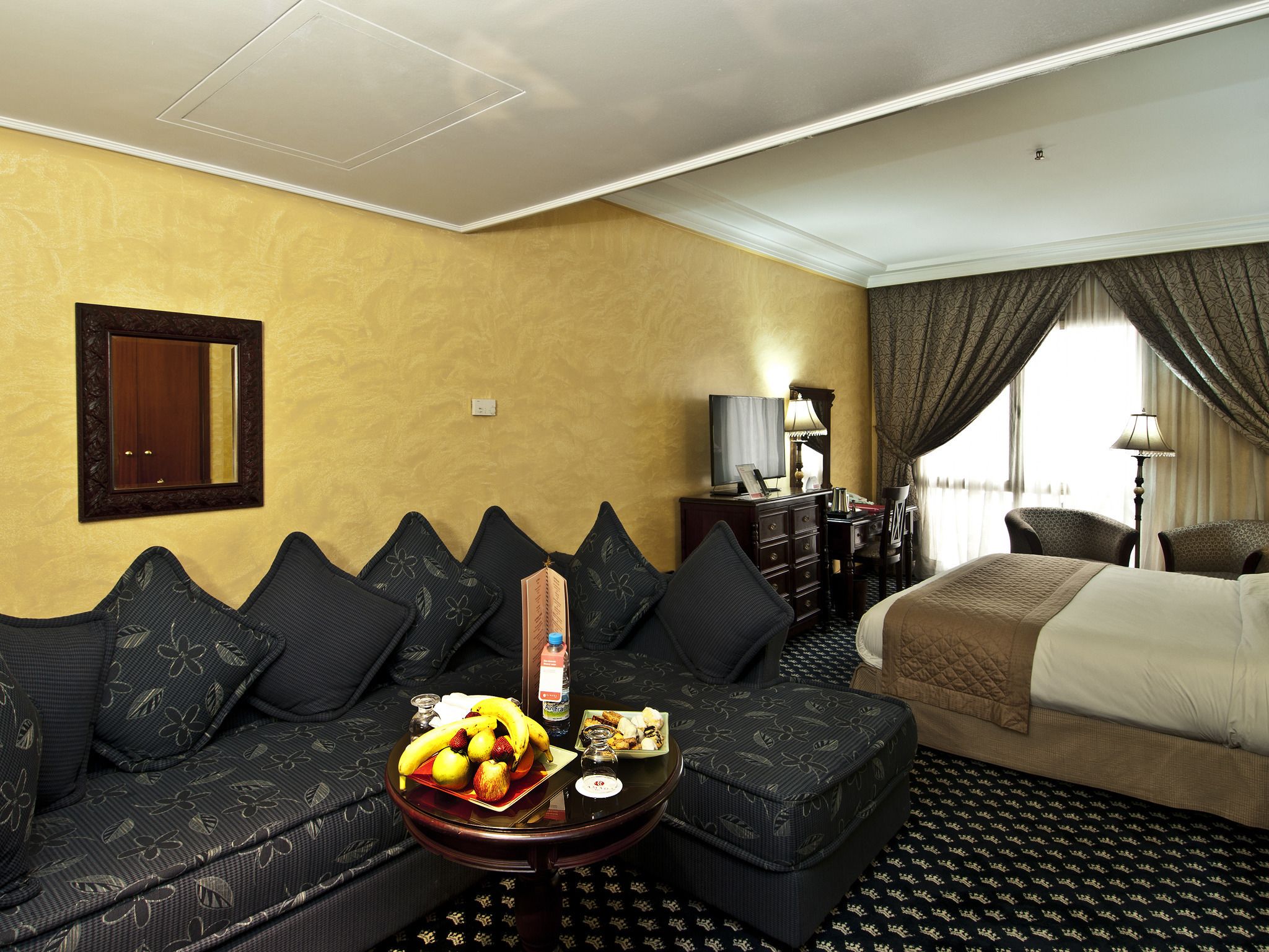 Habitación de hotel con sofá, una pequeña mesa con fruta, cama, escritorio con lámpara y espejo en la pared.