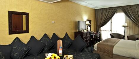 Habitación de hotel con sofá, una pequeña mesa con fruta, cama, escritorio con lámpara y espejo en la pared.