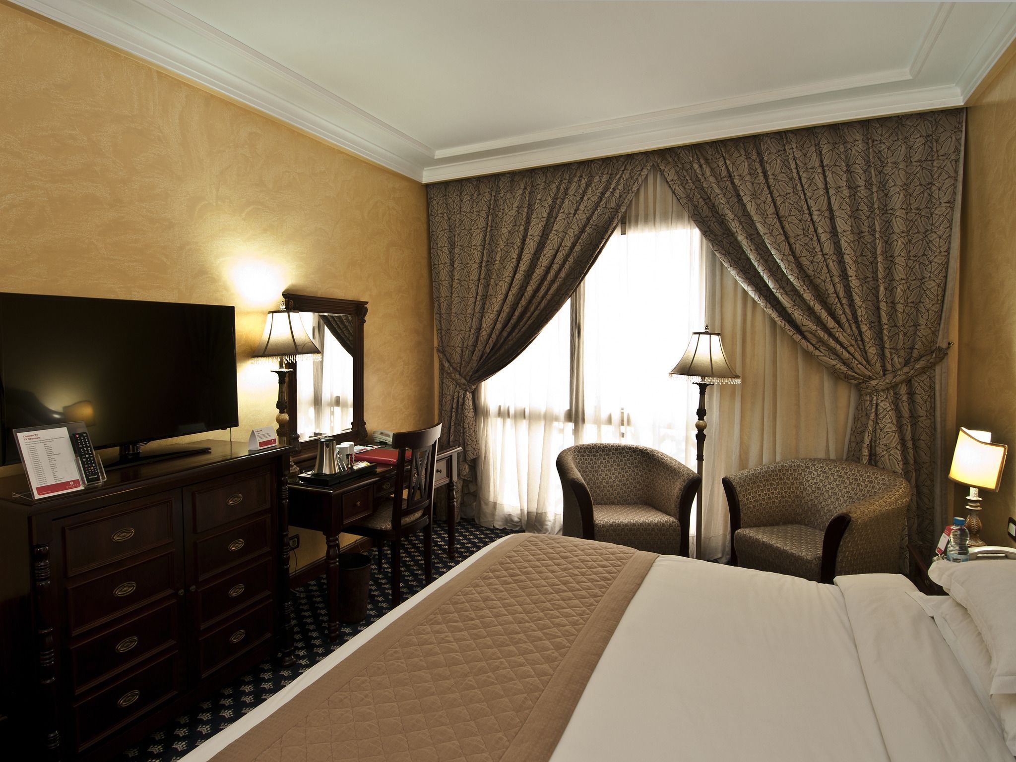 A hotel room with a bed, a desk with a television, two chairs, and a large window with curtains.