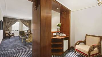 A hotel room with a wooden wardrobe, a small table with a vase, a chair, and a sofa.