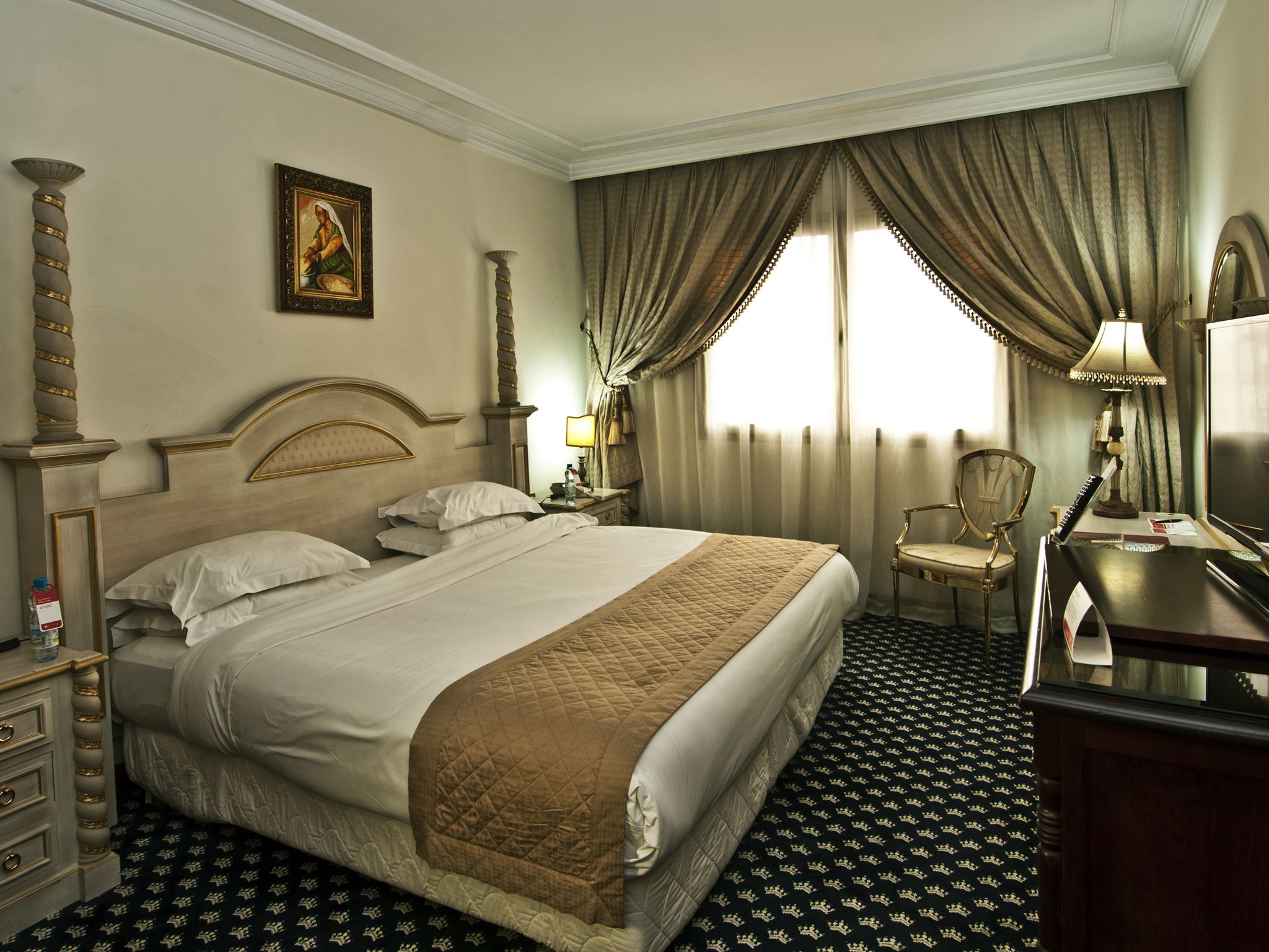 Une chambre d’hôtel avec un grand lit, un bureau avec une lampe, une chaise, une table de chevet, un tableau au mur et une fenêtre avec des rideaux.