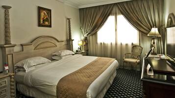 Une chambre d’hôtel avec un grand lit, un bureau avec une lampe, une chaise, une table de chevet, un tableau au mur et une fenêtre avec des rideaux.