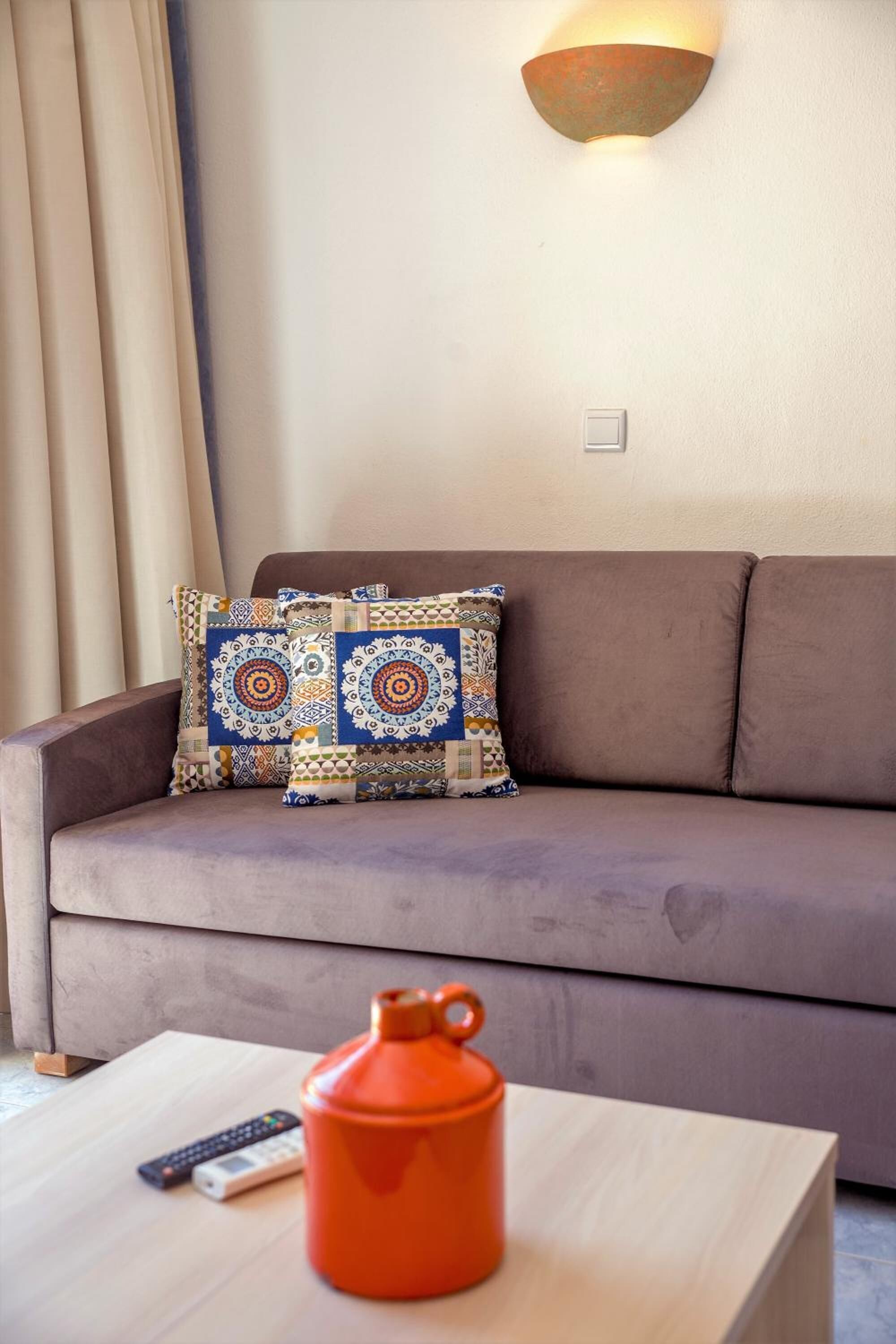 Ein Wohnzimmer mit einer Couch, gemusterten Kissen, einer Wandleuchte und einem Couchtisch mit einem roten Behälter und Fernbedienungen.