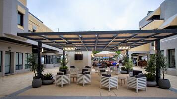 Een terras met tafels en stoelen, omringd door potplanten, onder een overdekte constructie.
