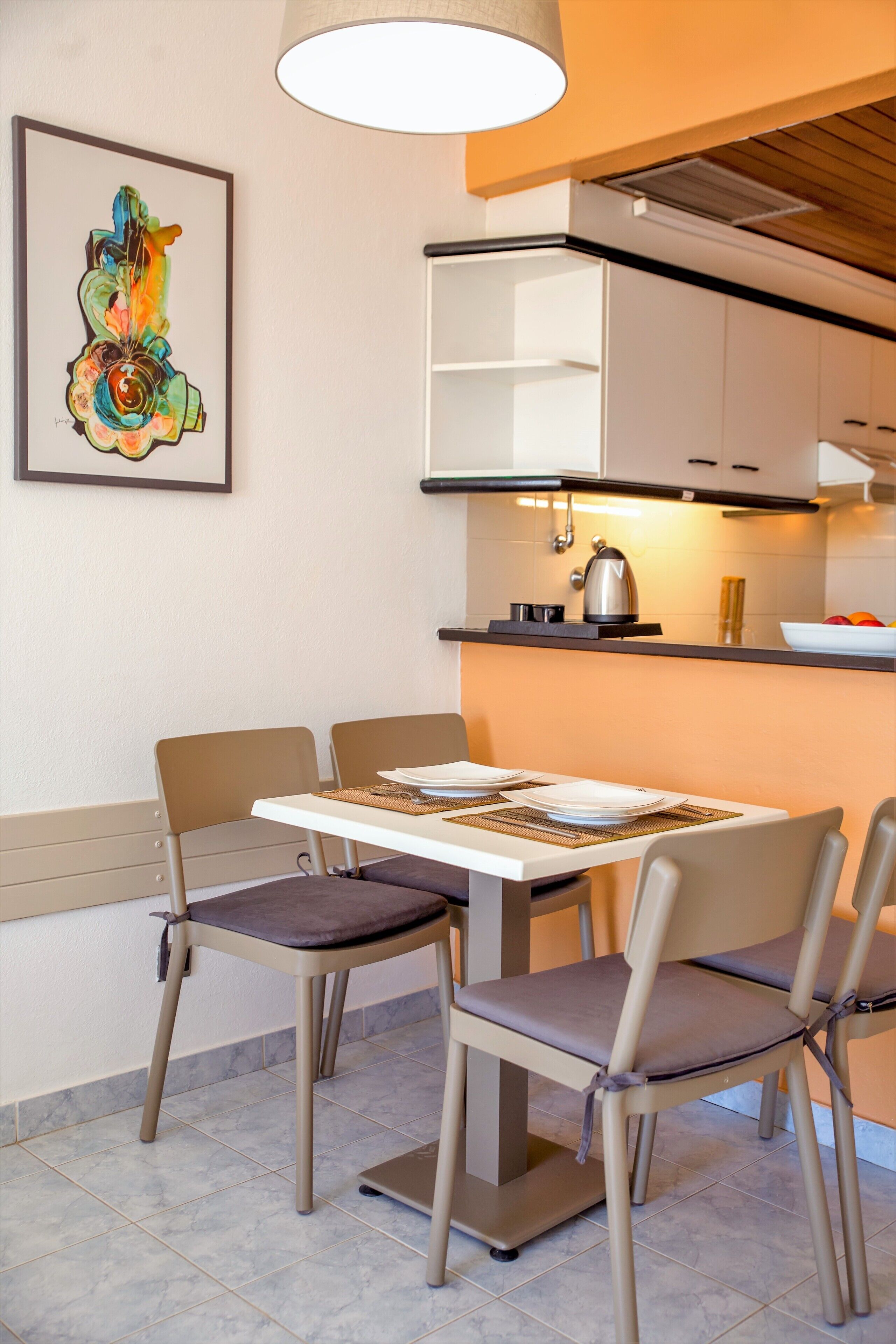 A modern kitchen with a dining table set for two, a kettle, and a bowl of fruit on the counter.