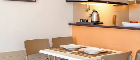 A modern kitchen with a dining table set for two, a kettle, and a bowl of fruit on the counter.