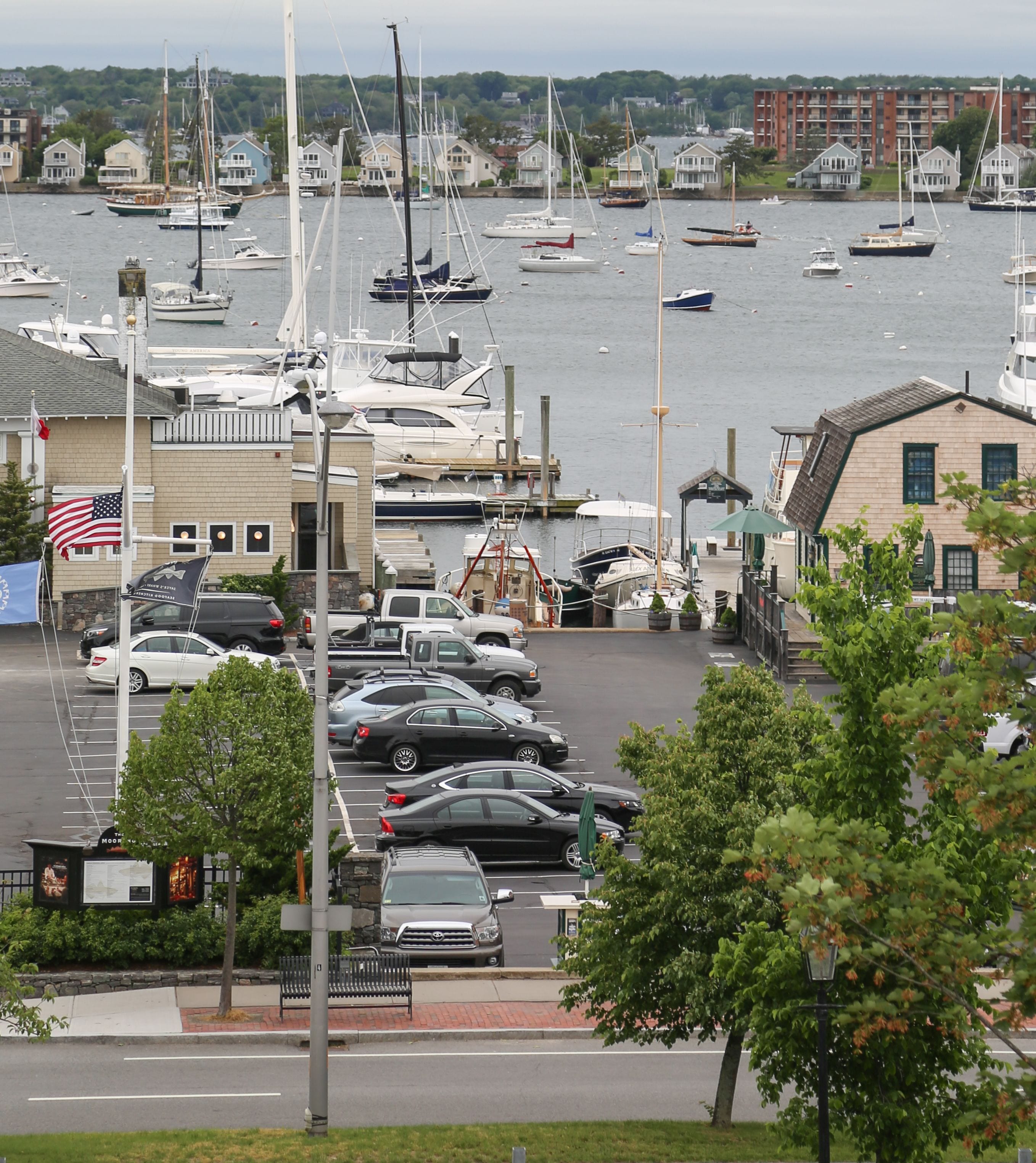 A marina with boats docked, a parking lot with cars, and buildings with American flags.