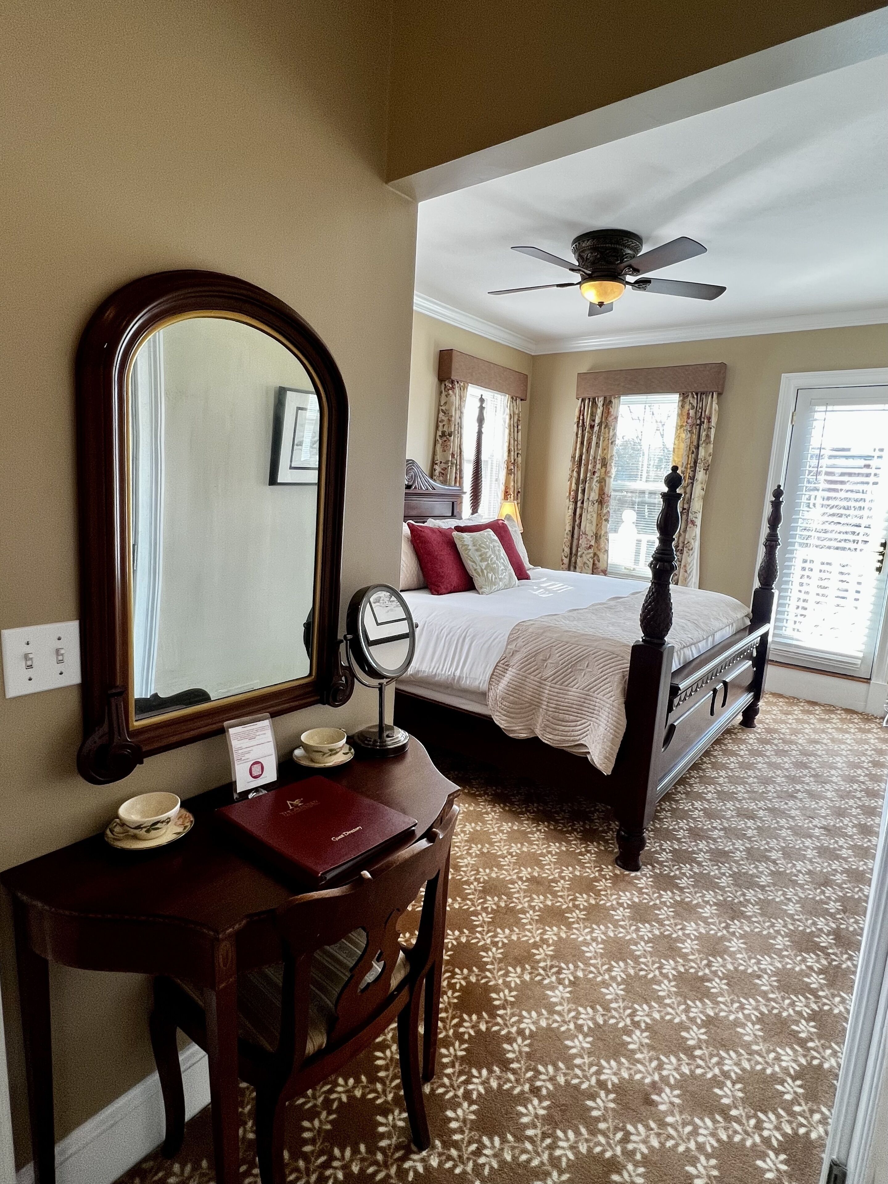 A hotel room with a large mirror, a wooden desk, a bed with a headboard, a ceiling fan, and a window with curtains.
