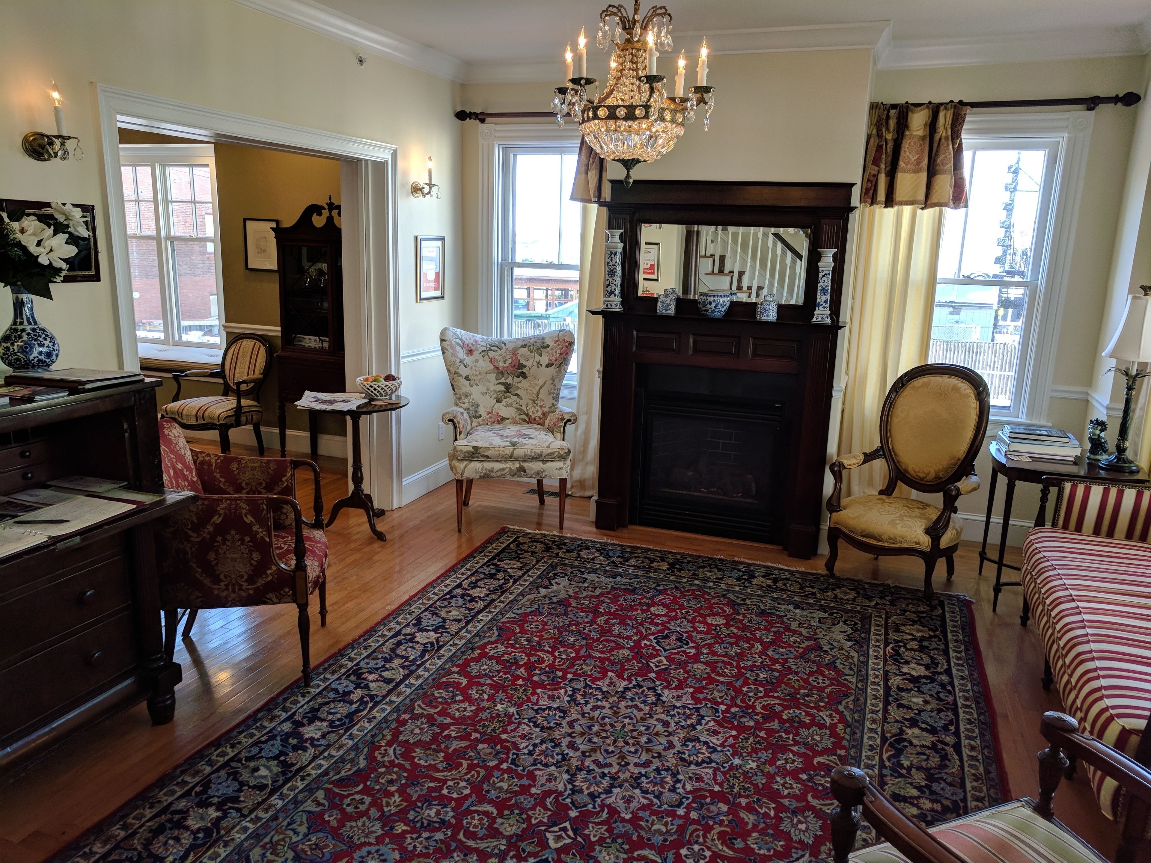 A traditional living room with a fireplace, floral armchairs, a large patterned rug, and a chandelier.
