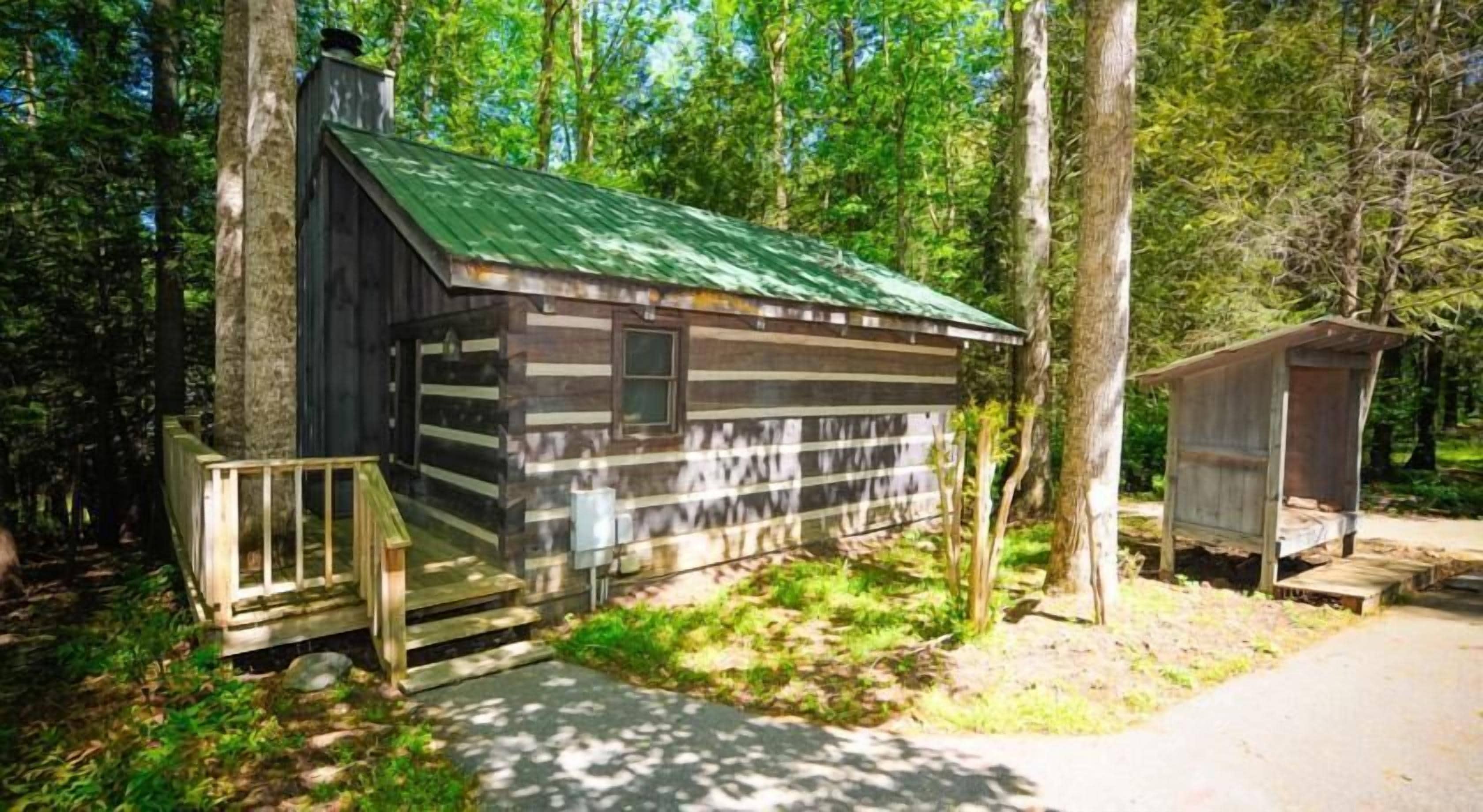 A rustic cabin with a green roof, a wooden deck, and a small shed in a wooded area.