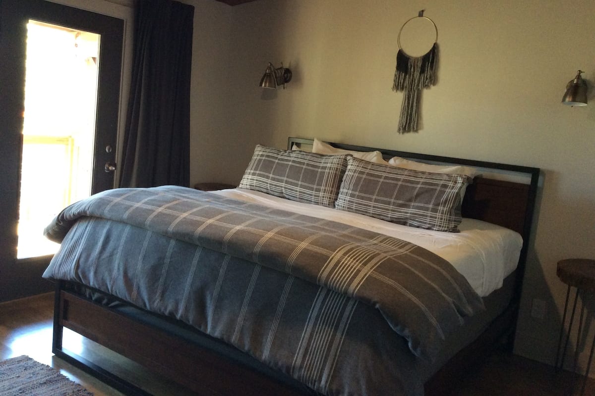 A bedroom with a bed, a door, a wall-mounted lamp, and a macrame wall hanging.