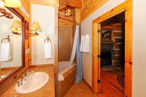 A wooden bathroom with a sink, mirror, and bathtub. There are towels hanging on the wall and a painting above the bathtub.