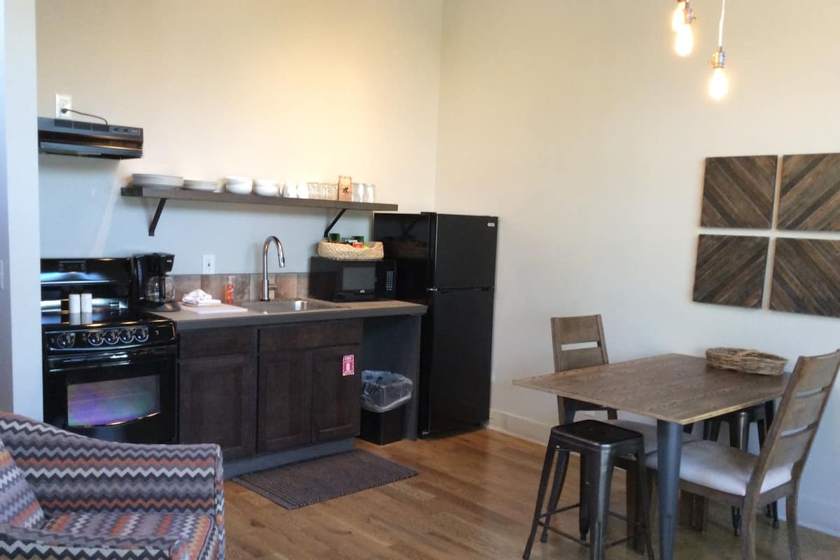 A compact kitchen with a stove, sink, and refrigerator, a dining table with chairs, and a patterned armchair.