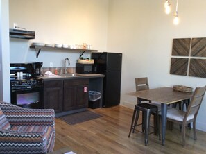 A compact kitchen with a stove, sink, and refrigerator, a dining table with chairs, and a patterned armchair.