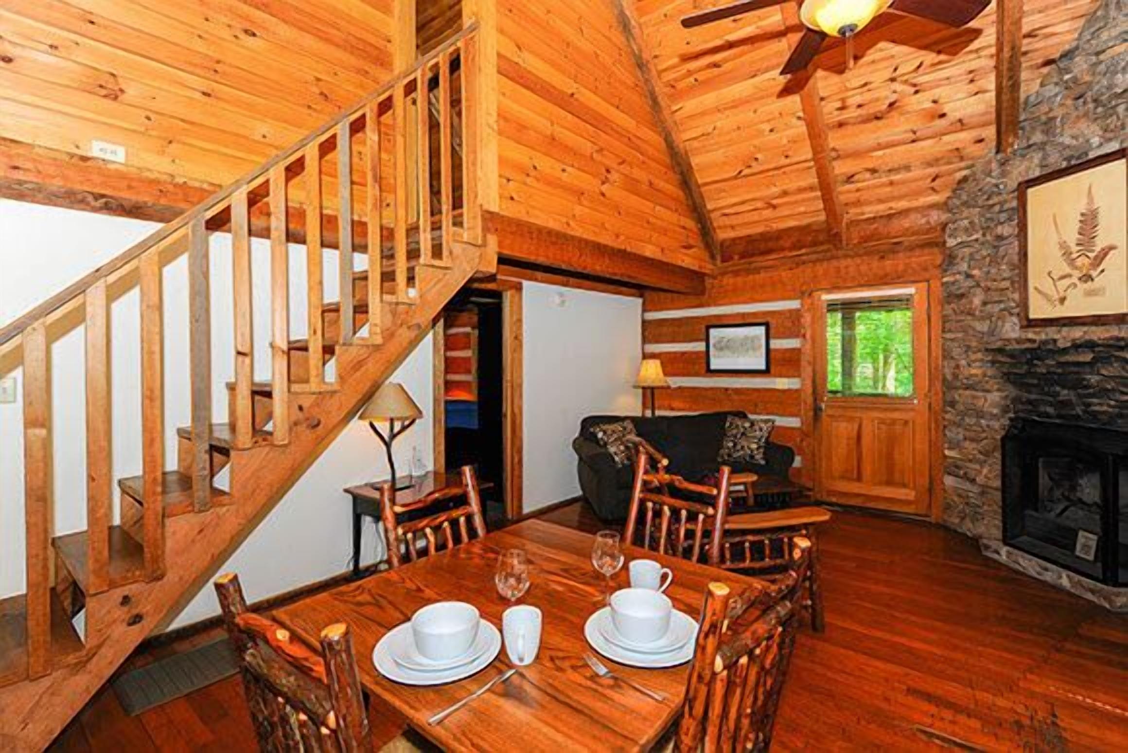 A wooden cabin interior with a dining area, a fireplace, and a staircase.