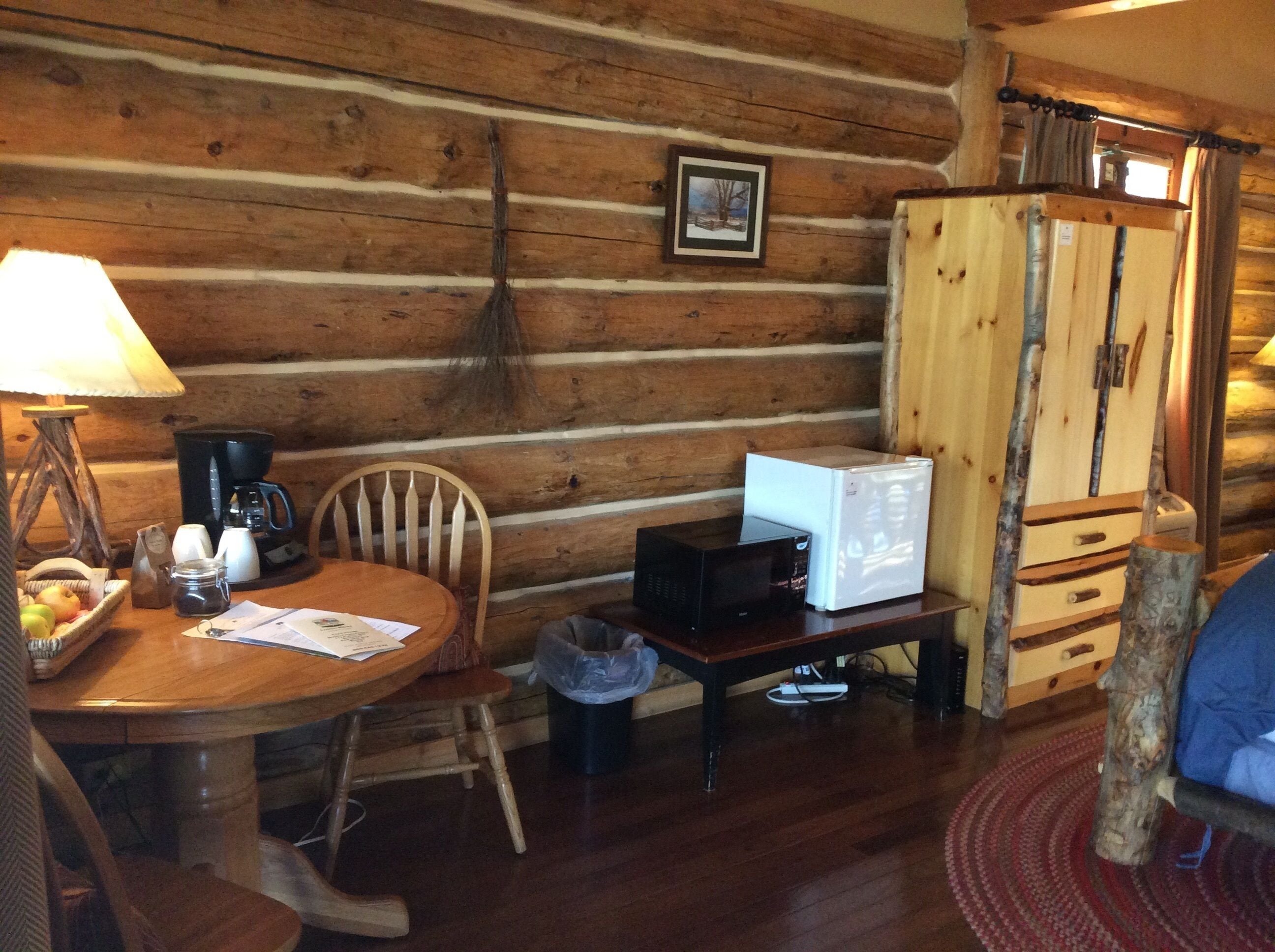 A cozy cabin interior with a wooden dining table, chairs, a lamp, a coffee maker, a microwave, and a wooden cabinet.