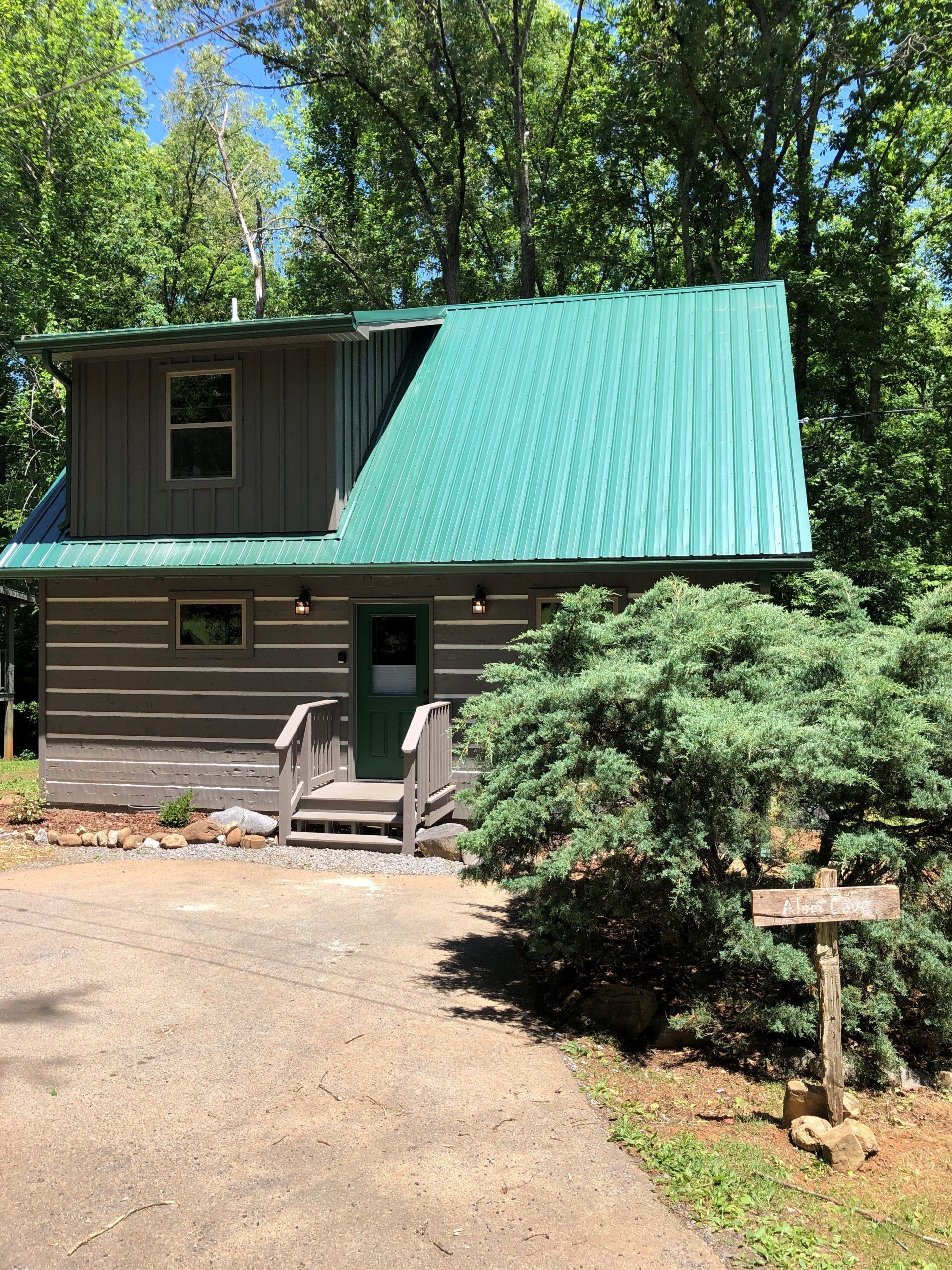 A two-story lodge with a green metal roof, a front porch with steps, and a sign reading "Alm Cove".