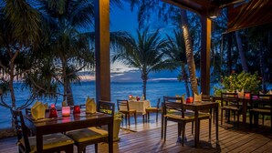 An outdoor dining area with wooden tables and chairs, overlooking a body of water with palm trees. - TUI BLUE The Passage Samui Pool Villas with Private Beach Resort (Koh Samui)