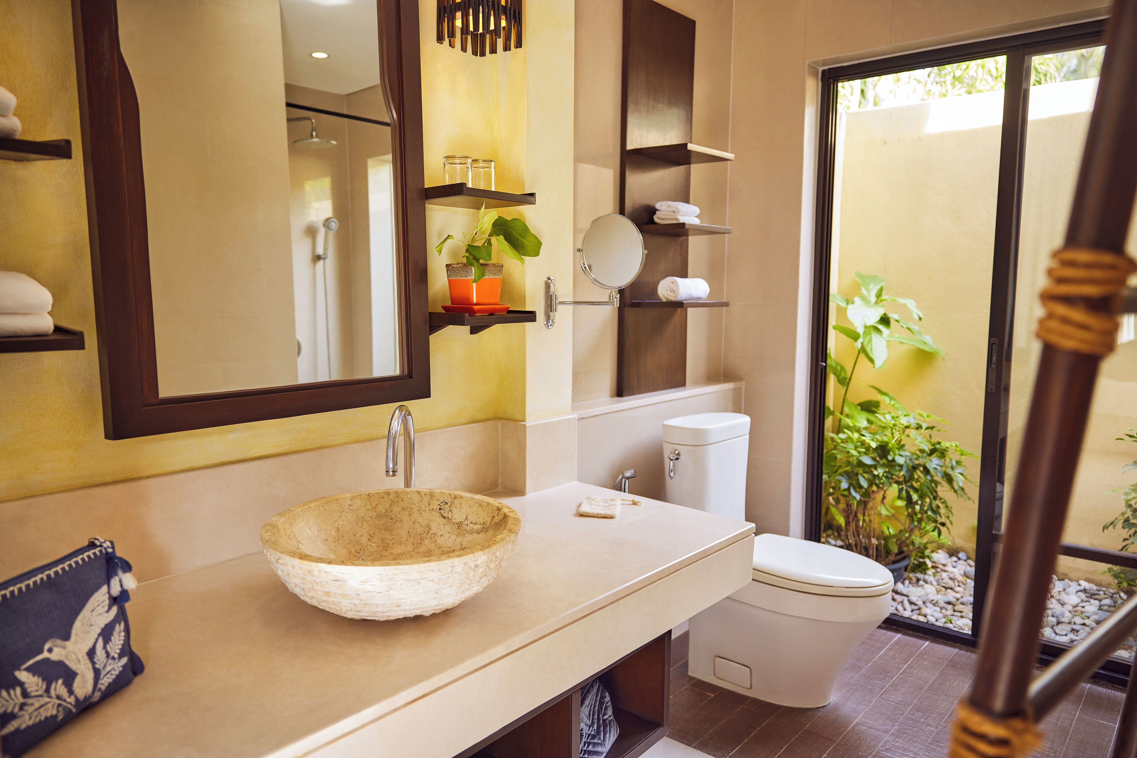 A bathroom with a large bowl sink, a toilet, and a mirror. There are shelves with towels and a potted plant.