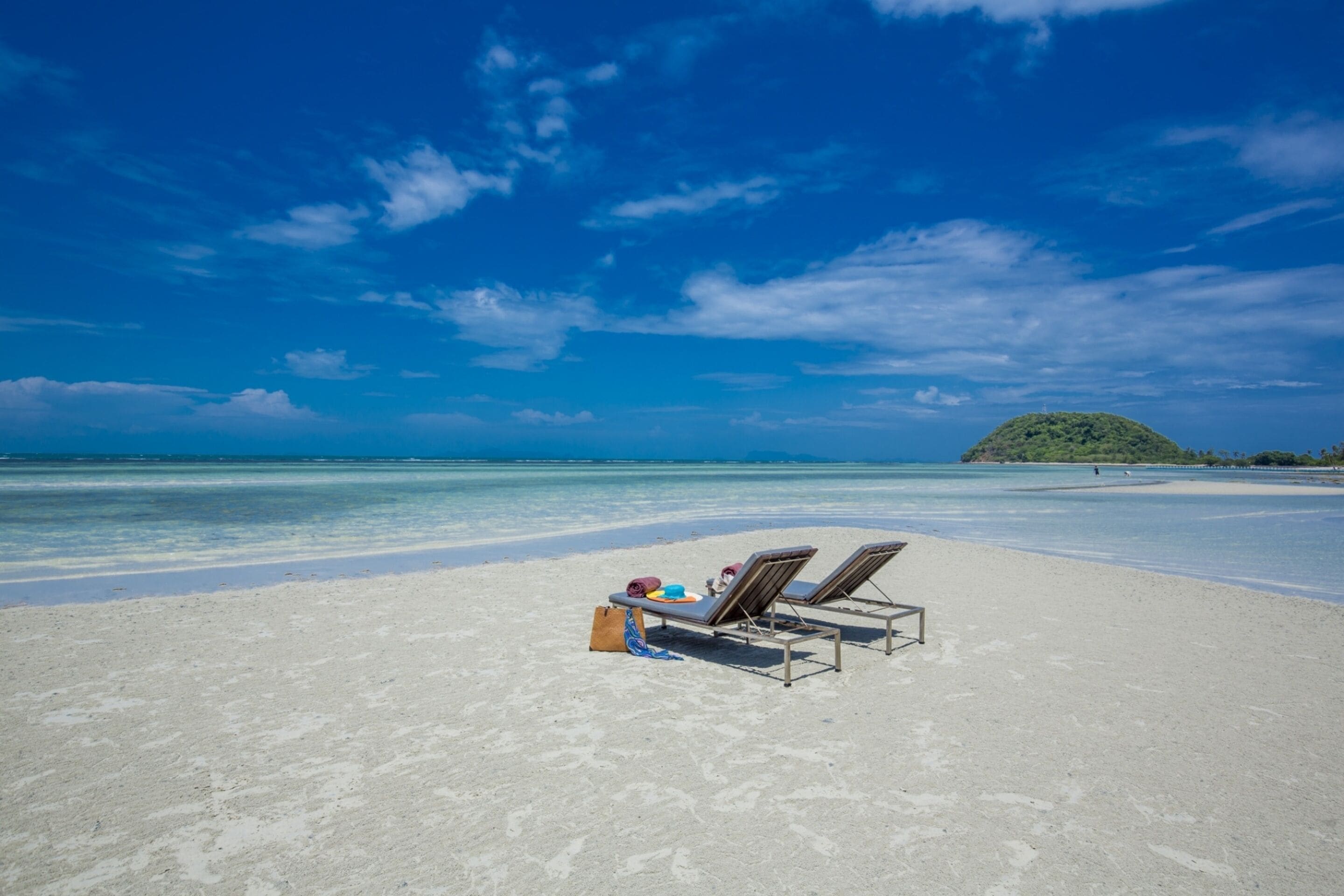 A beach with two sun loungers and a bag on the sand, a clear blue sky, and a distant island.