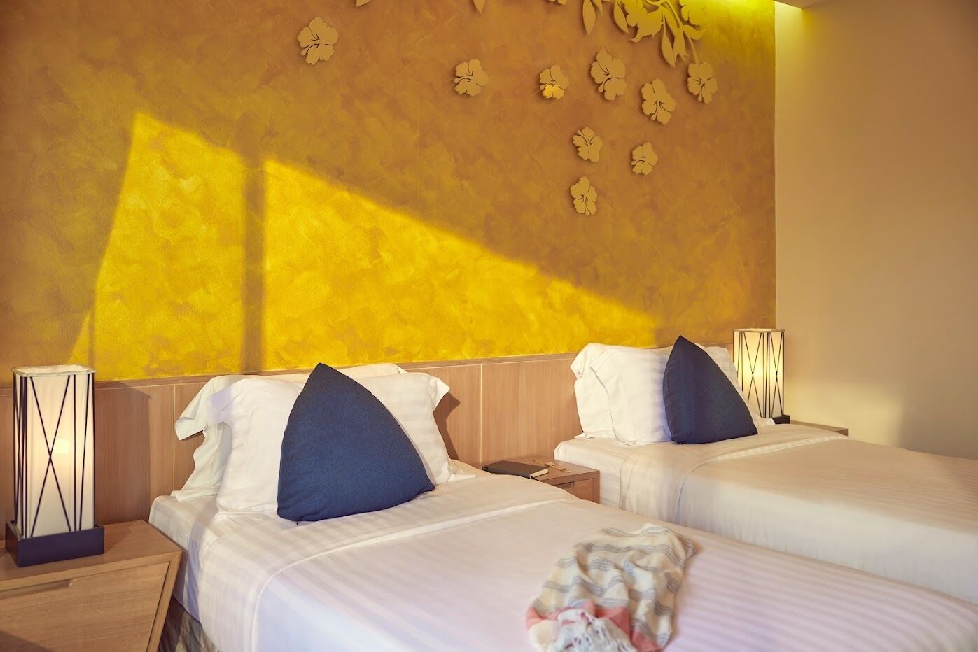 Une chambre d’hôtel avec deux lits, chacun agrémenté de draps blancs et d’oreillers bleu foncé. Un mur décoratif orné d’un motif floral et un luminaire se trouvent sur le côté gauche.