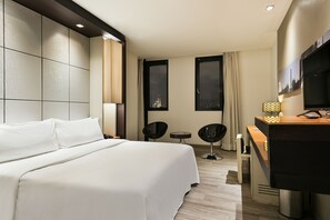 A modern hotel room with a large bed, a desk with a flat-screen TV, two chairs, and a small table.