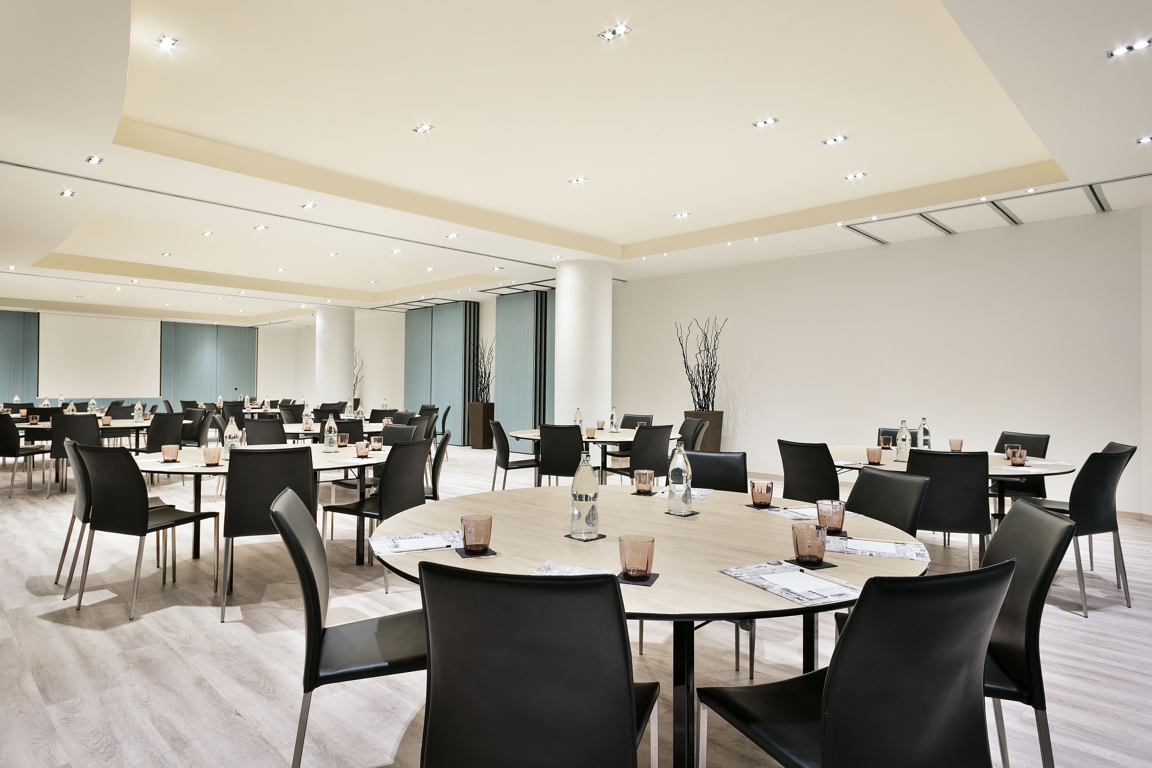 Een ruime vergaderzaal met meerdere tafels en stoelen, voorzien van glaswerk en documenten, en een modern plafondontwerp.
