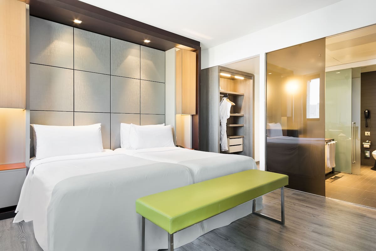 Une chambre d’hôtel moderne dotée d’un grand lit, d’une banquette, d’un dressing et d’une salle de bain avec une douche en verre.