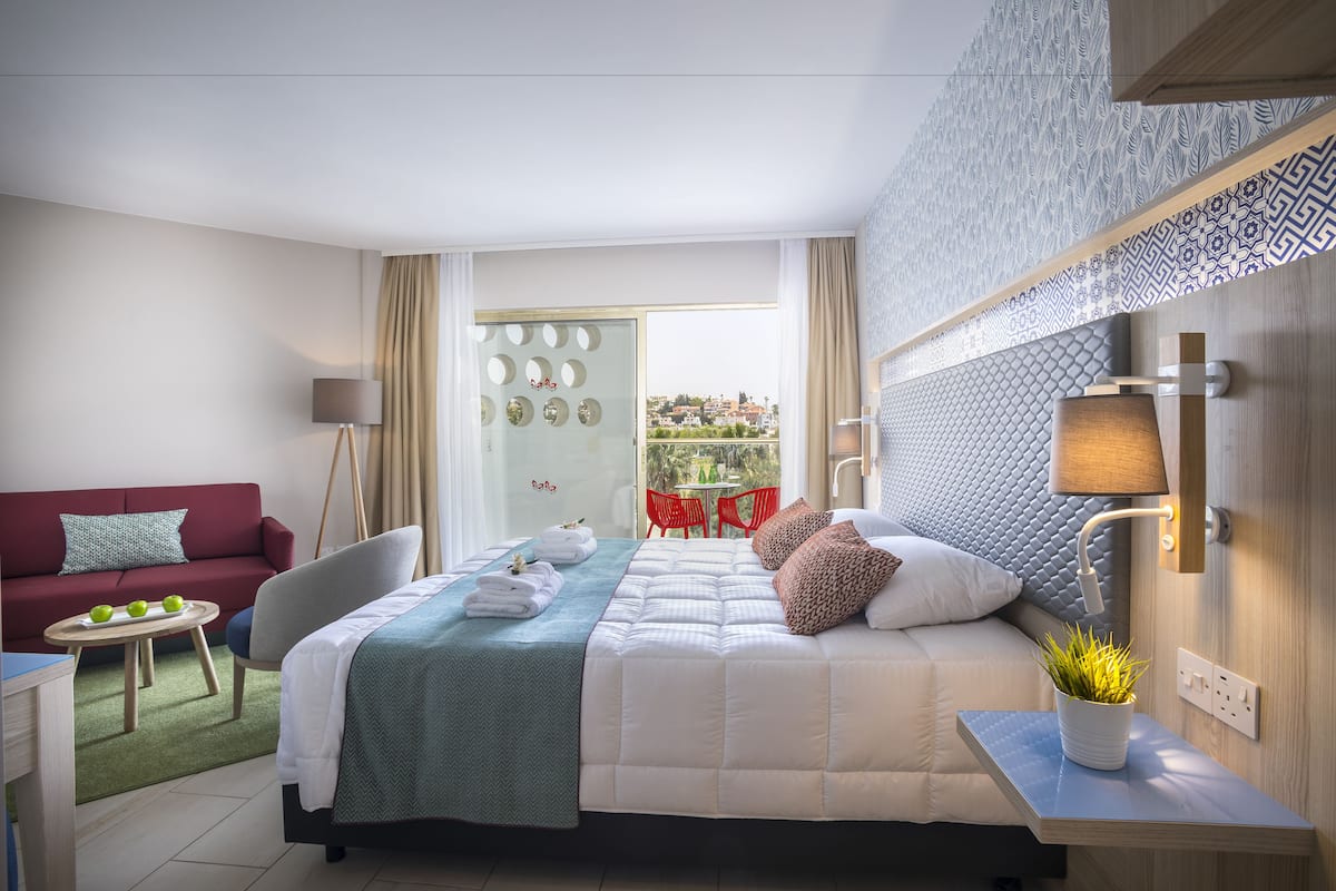 Une chambre d’hôtel avec un lit, un canapé, une petite table avec des pommes, une plante en pot et une vue sur l’extérieur.