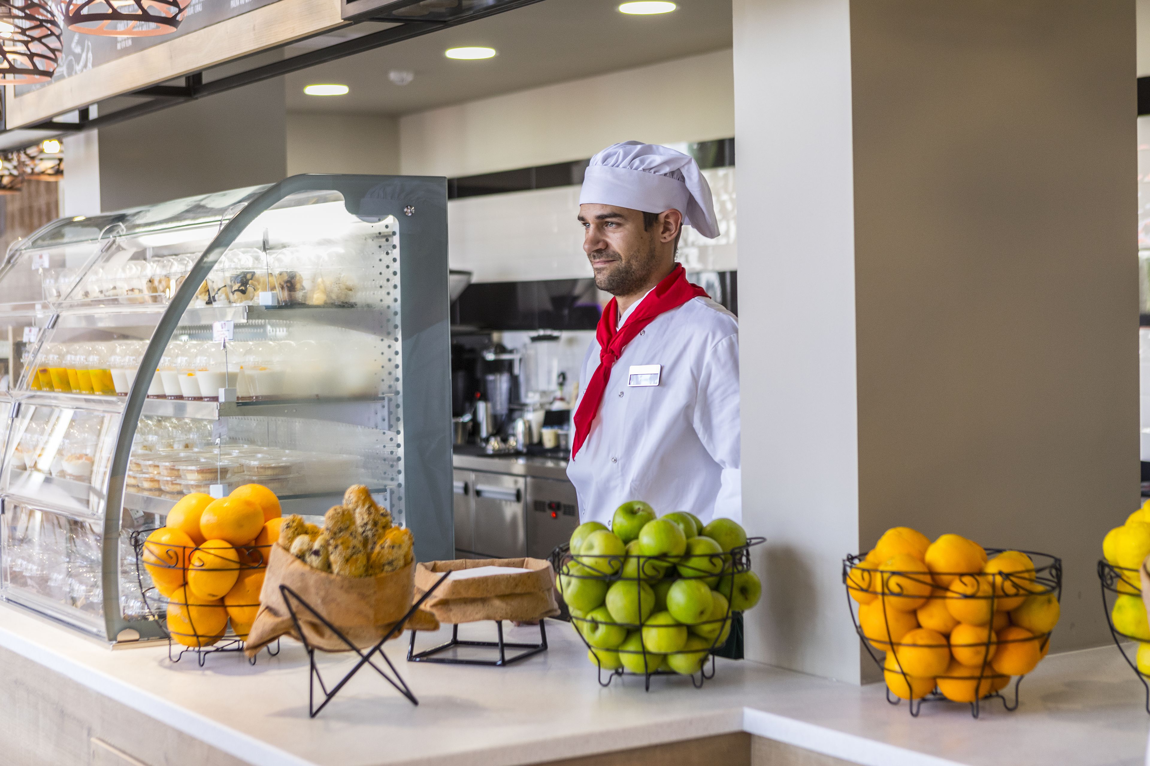 A chef in a white uniform with a red scarf standing behind a counter with fresh fruits and pastries.