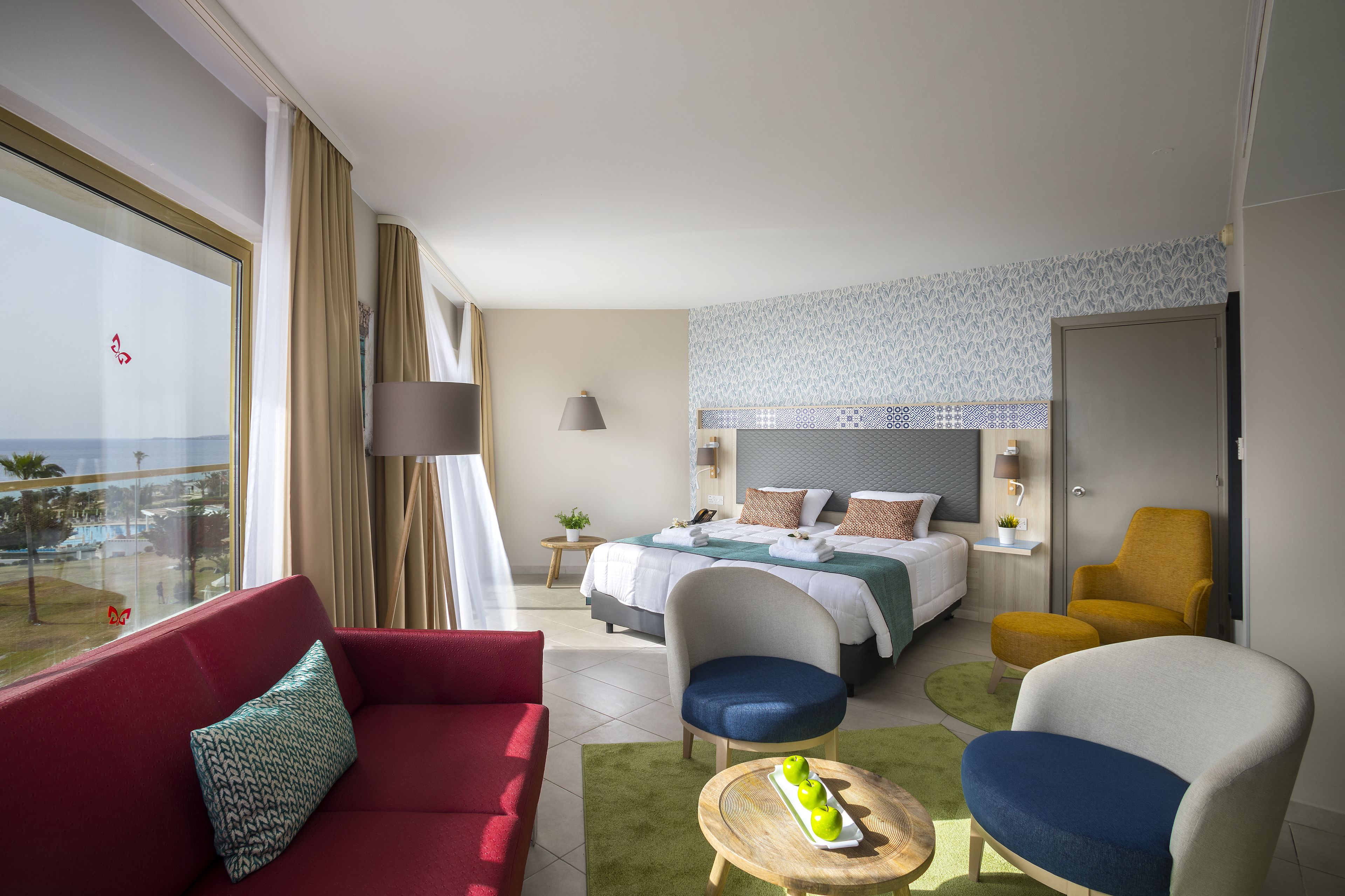 Una camera d'albergo con un letto, un divano rosso, due poltrone, una gialla e una blu, e un tavolino con delle mele verdi.