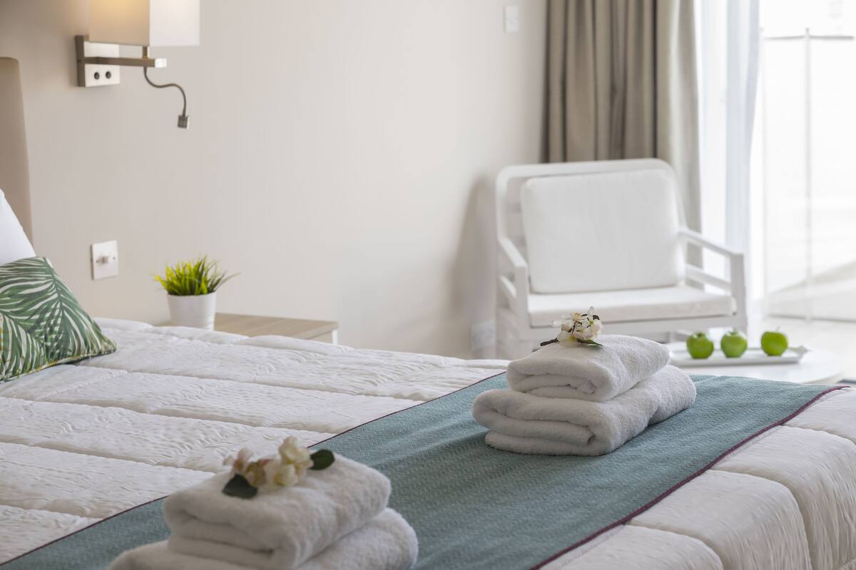 Un lit bien fait, avec du linge de lit blanc et un plaid vert, une chaise blanche, une petite plante en pot et un plateau avec des pommes vertes.