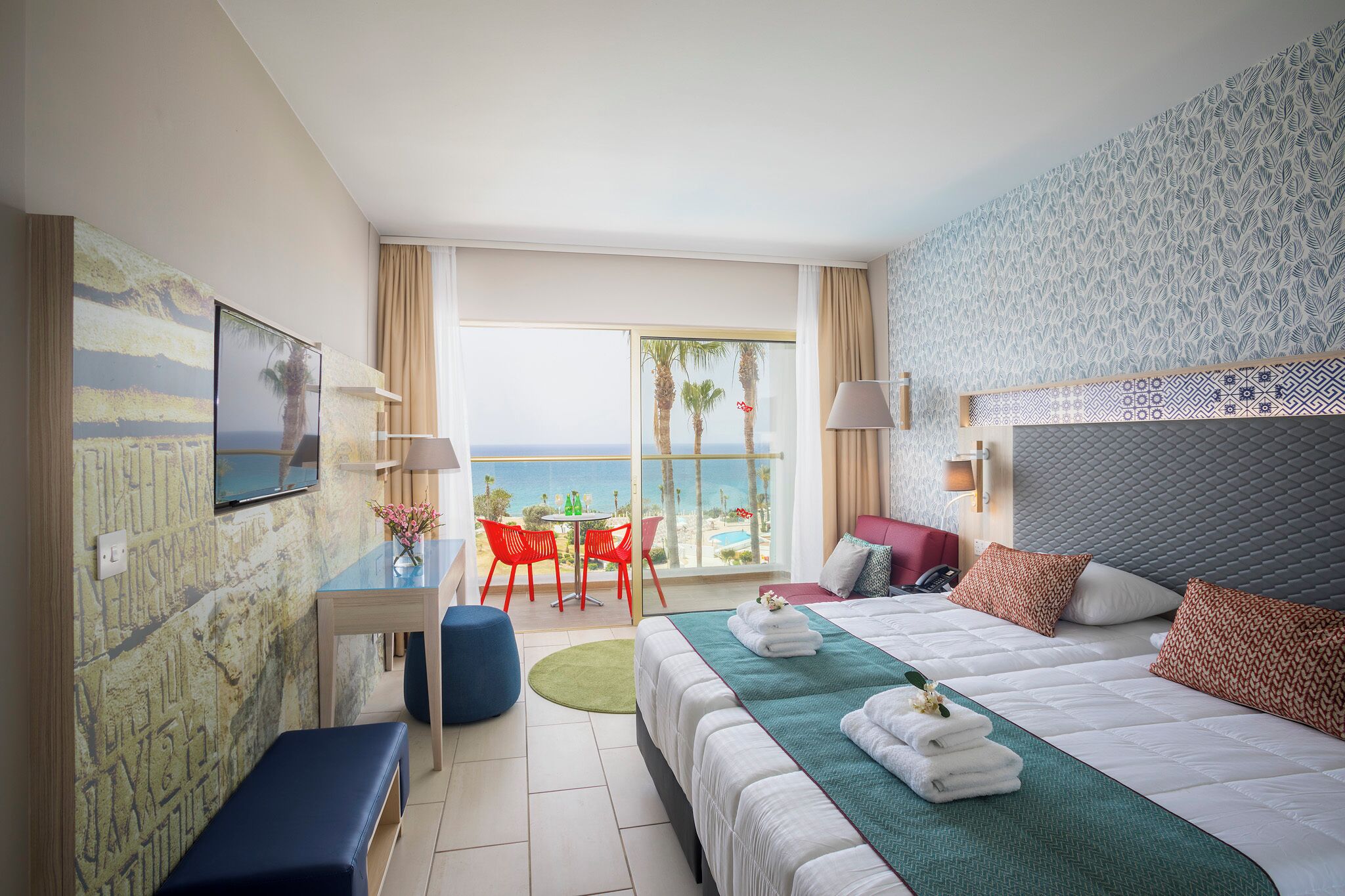 Ein Hotelzimmer mit Bett, Schreibtisch, Stuhl, einem kleinen Tisch, einer Bank und Blick aufs Meer.