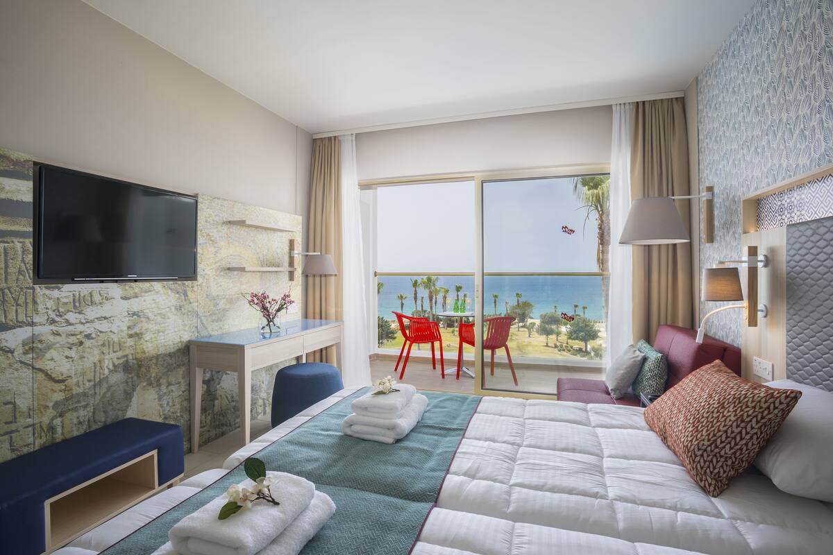 Une chambre d’hôtel avec un lit, un bureau, une télévision et un balcon doté de chaises rouges et offrant une vue sur l’océan.