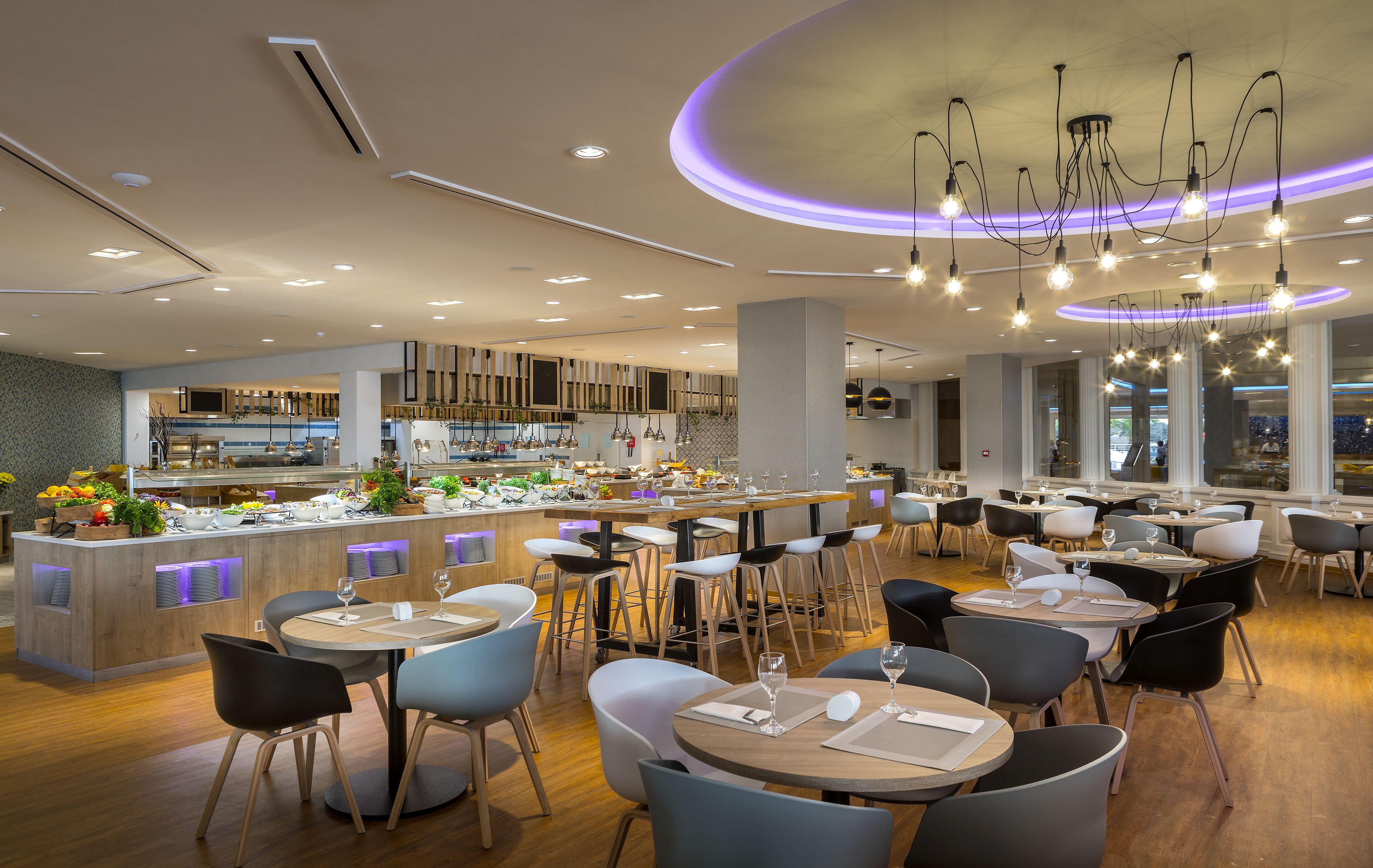 A modern restaurant with a central bar area, multiple dining tables, and contemporary lighting fixtures.
