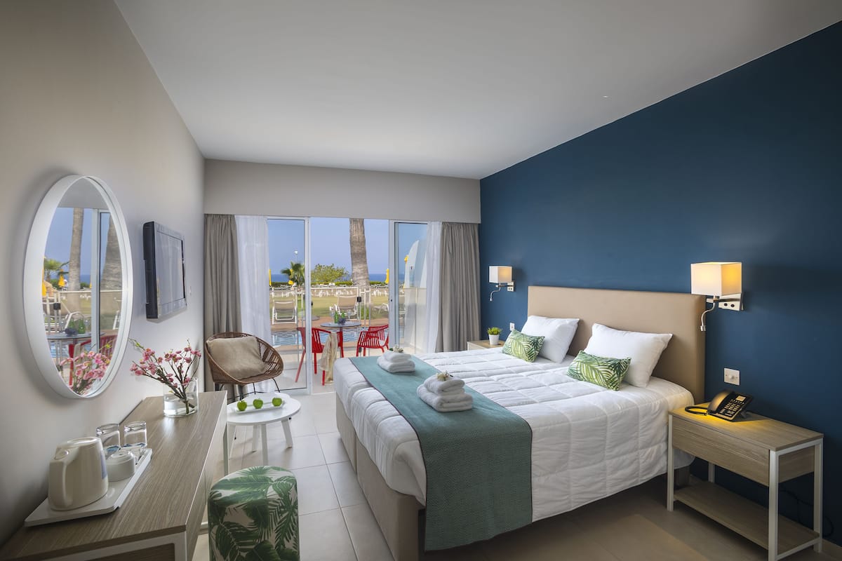 Une chambre d’hôtel moderne avec un grand lit, un bureau avec une chaise, une petite table ornée d’un vase de fleurs et une vue sur l’extérieur.