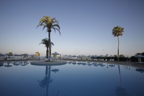 Piscina cercada por palmeiras, cadeiras de praia e guarda-sóis.