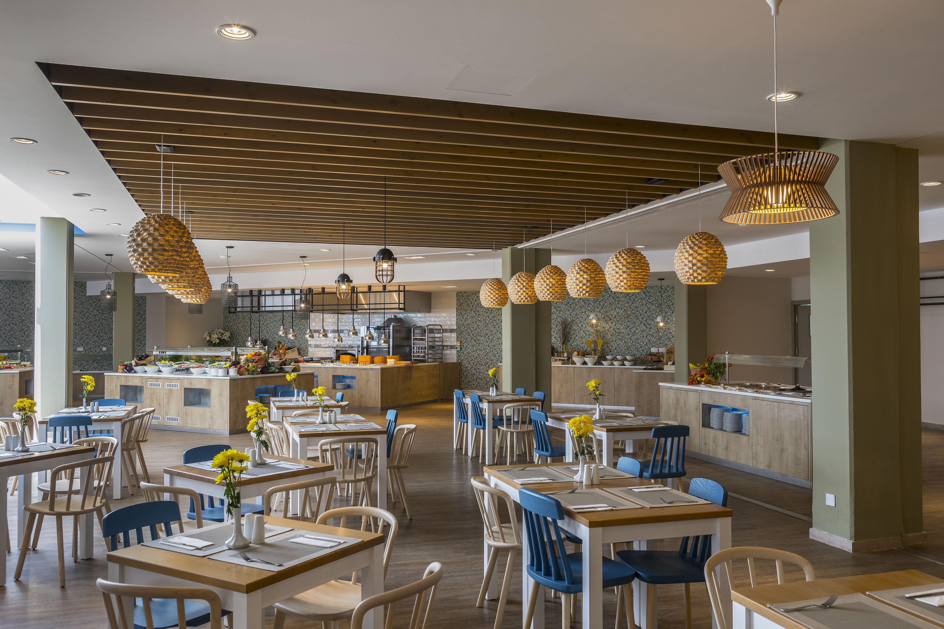 Un comedor amplio con varias mesas y sillas, una zona de cocina central y lámparas colgantes.