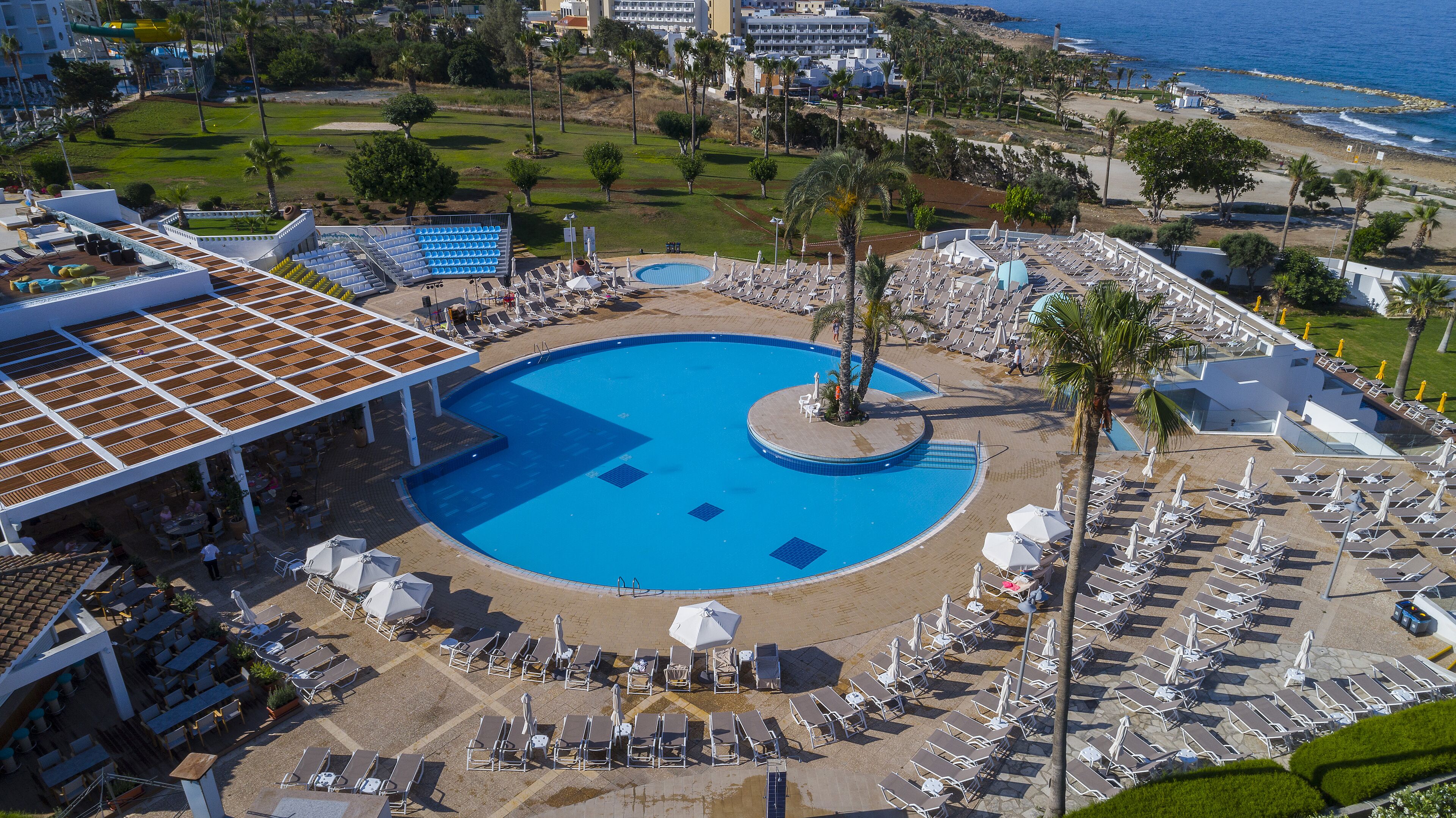 Une grande piscine entourée d’un espace aménagé avec des palmiers, des transats et des parasols, offrant une vue sur la mer.
