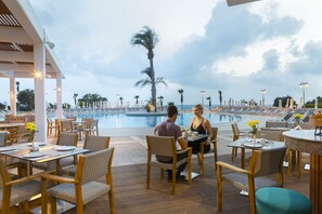 An outdoor dining area with tables and chairs, overlooking a pool and palm trees. - Leonardo Laura Beach & Splash Resort (Chlorakas)