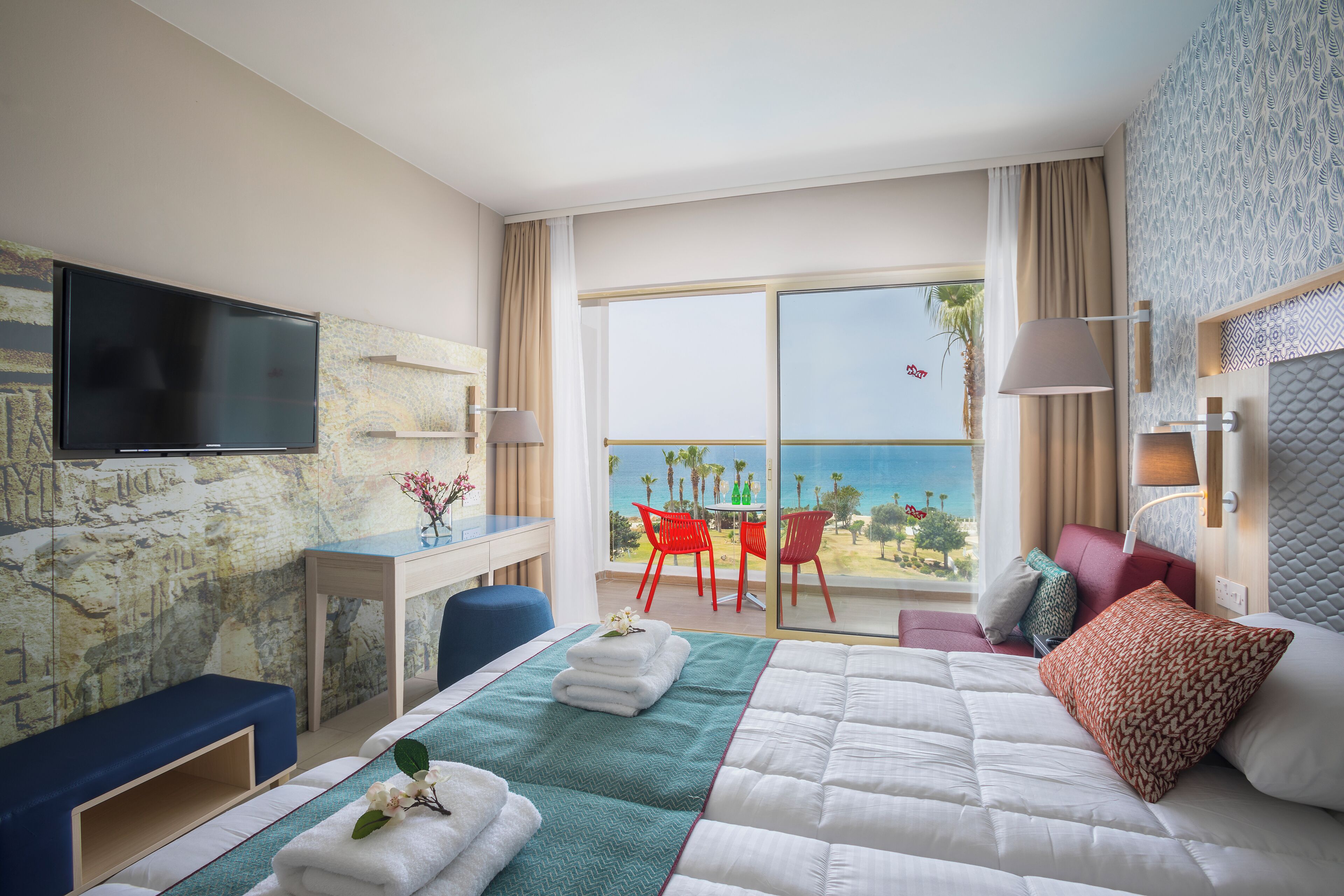 Ein Hotelzimmer mit Bett, Schreibtisch, Fernseher und einem Balkon mit roten Stühlen sowie Meerblick.