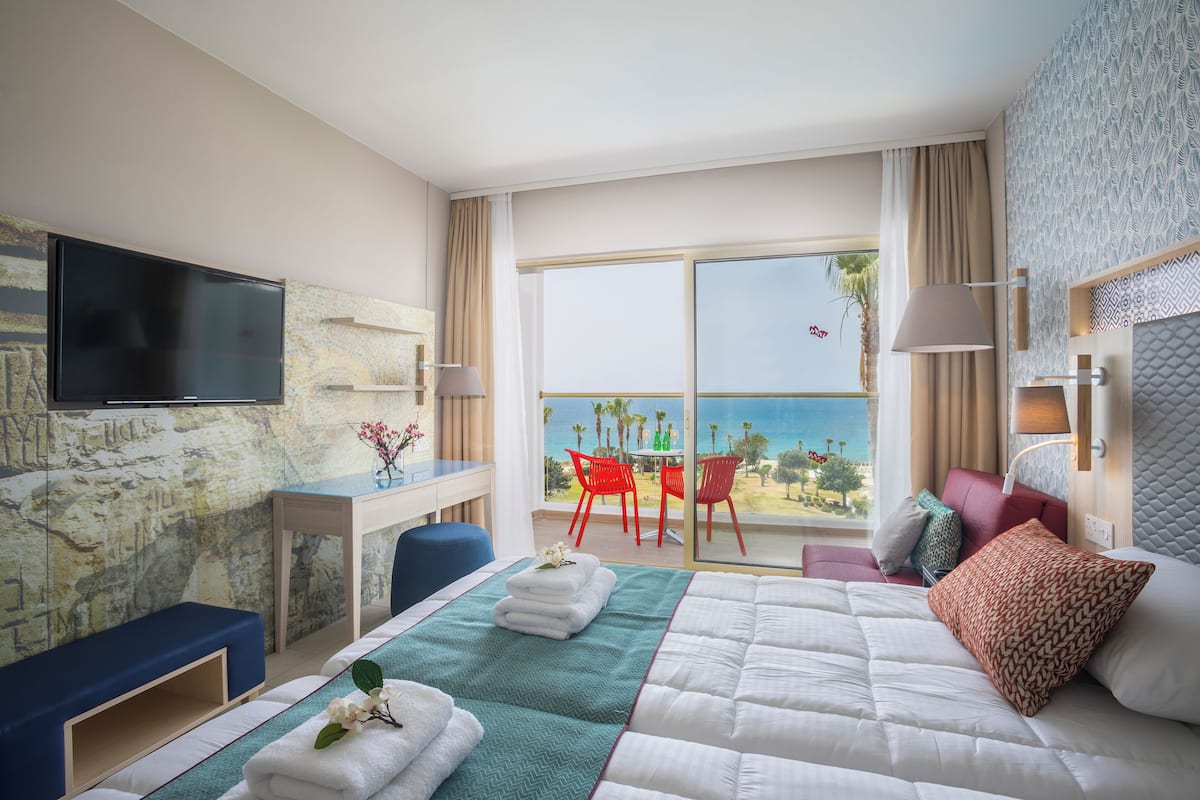 Une chambre d’hôtel avec un lit, un bureau, une télévision et un balcon doté de chaises rouges et offrant une vue sur l’océan.