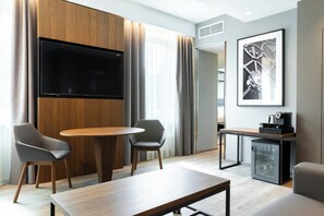A modern hotel room with a wooden dining table, two grey chairs, a flat-screen TV mounted on a wooden panel, a small refrigerator, and a framed abstract artwork on the wall. - AC Hotel by Marriott Riga (Riga)