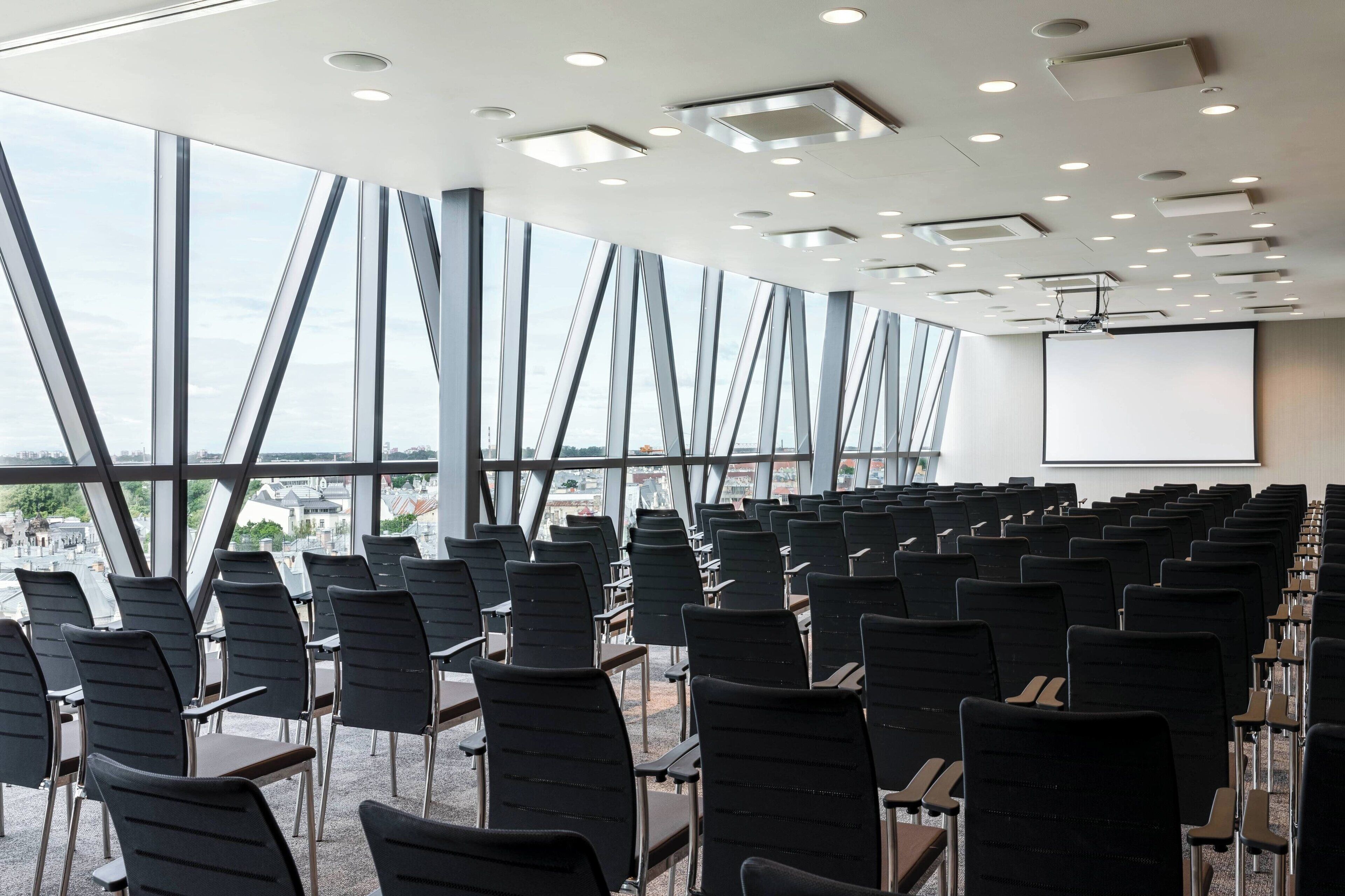 Un salón de conferencias con filas de sillas y mesas, ventanas amplias y una pantalla para proyector.