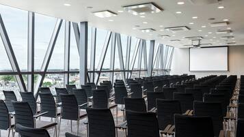 Un salón de conferencias con filas de sillas y mesas, ventanas amplias y una pantalla para proyector.