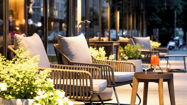 Wicker chairs with cushions, a table with a drink, and potted plants in an outdoor seating area with a city view. - AC Hotel by Marriott Riga (Riga)