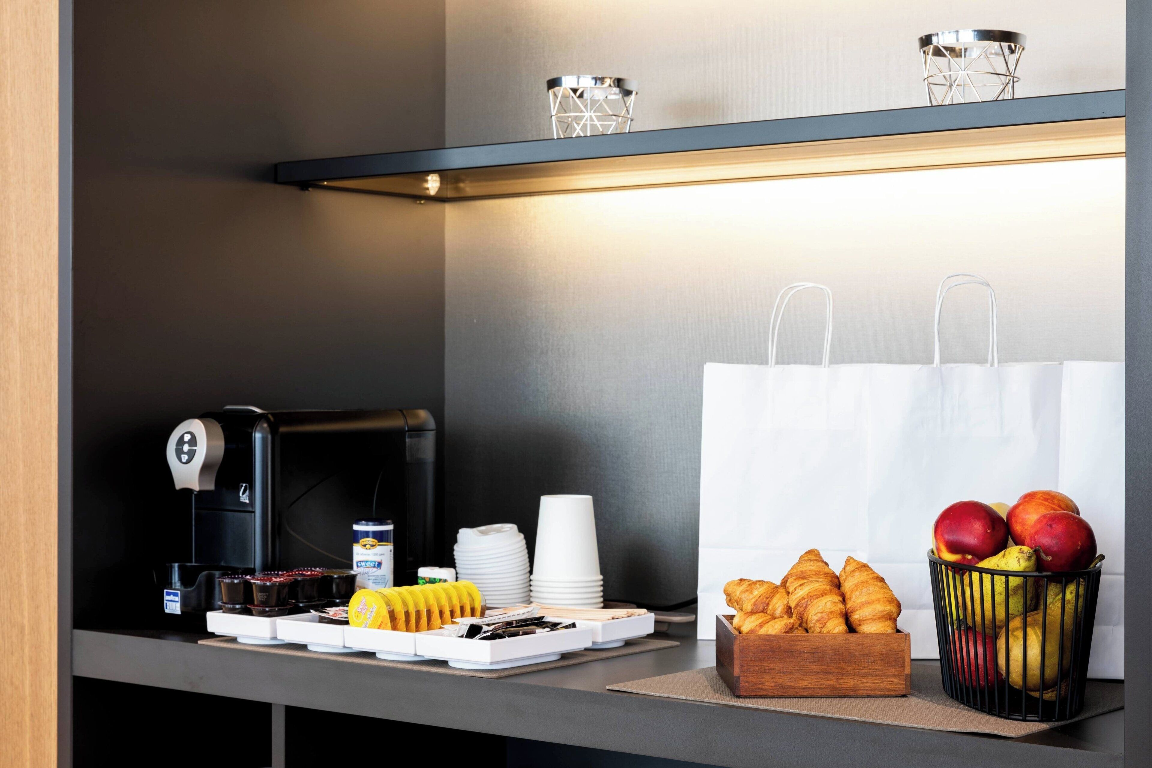 A breakfast setup with a coffee machine, croissants, fruits, and paper cups.