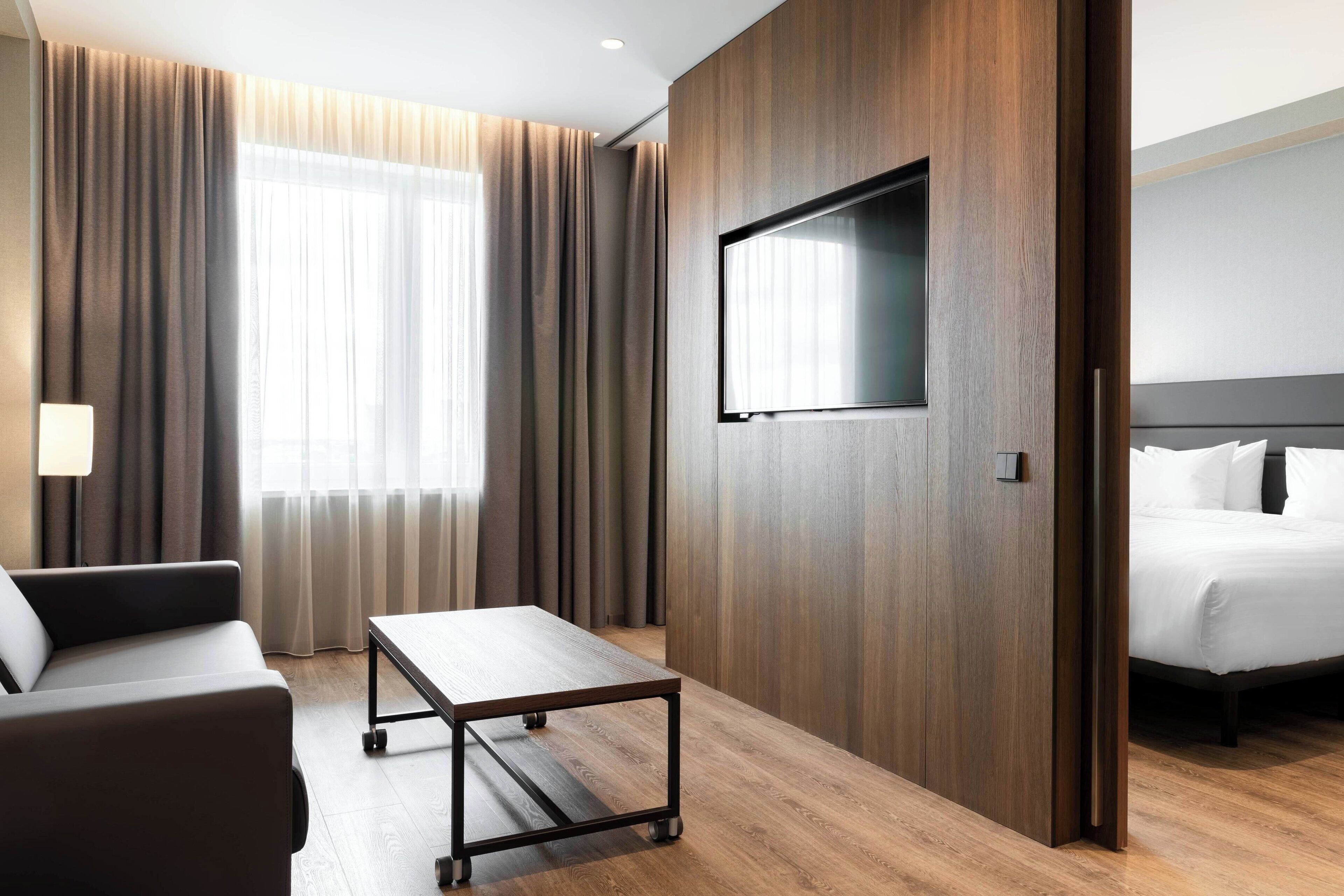 Una habitación de hotel moderna con suelo de madera, un sofá, una mesita, una cama y un televisor fijado a la pared.